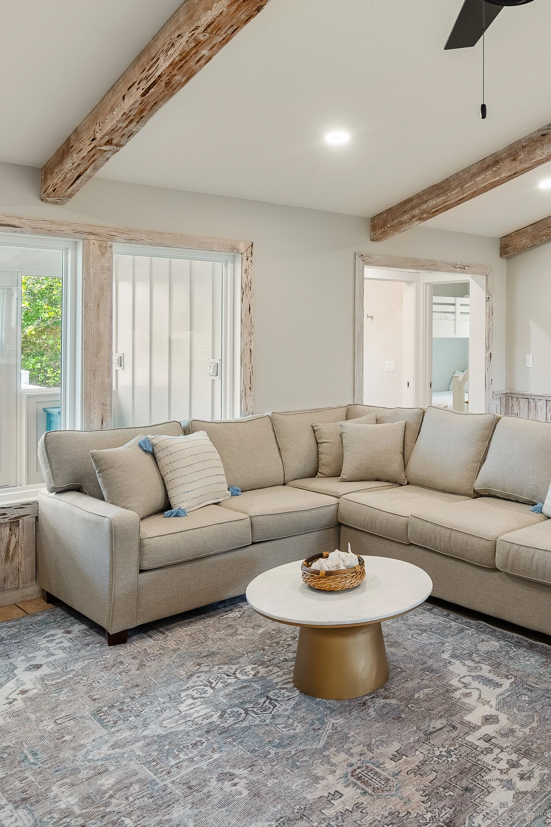 Living room with beige sectional sofa, decorative pillows, round coffee table, patterned rug, large windows, and exposed wooden beams.