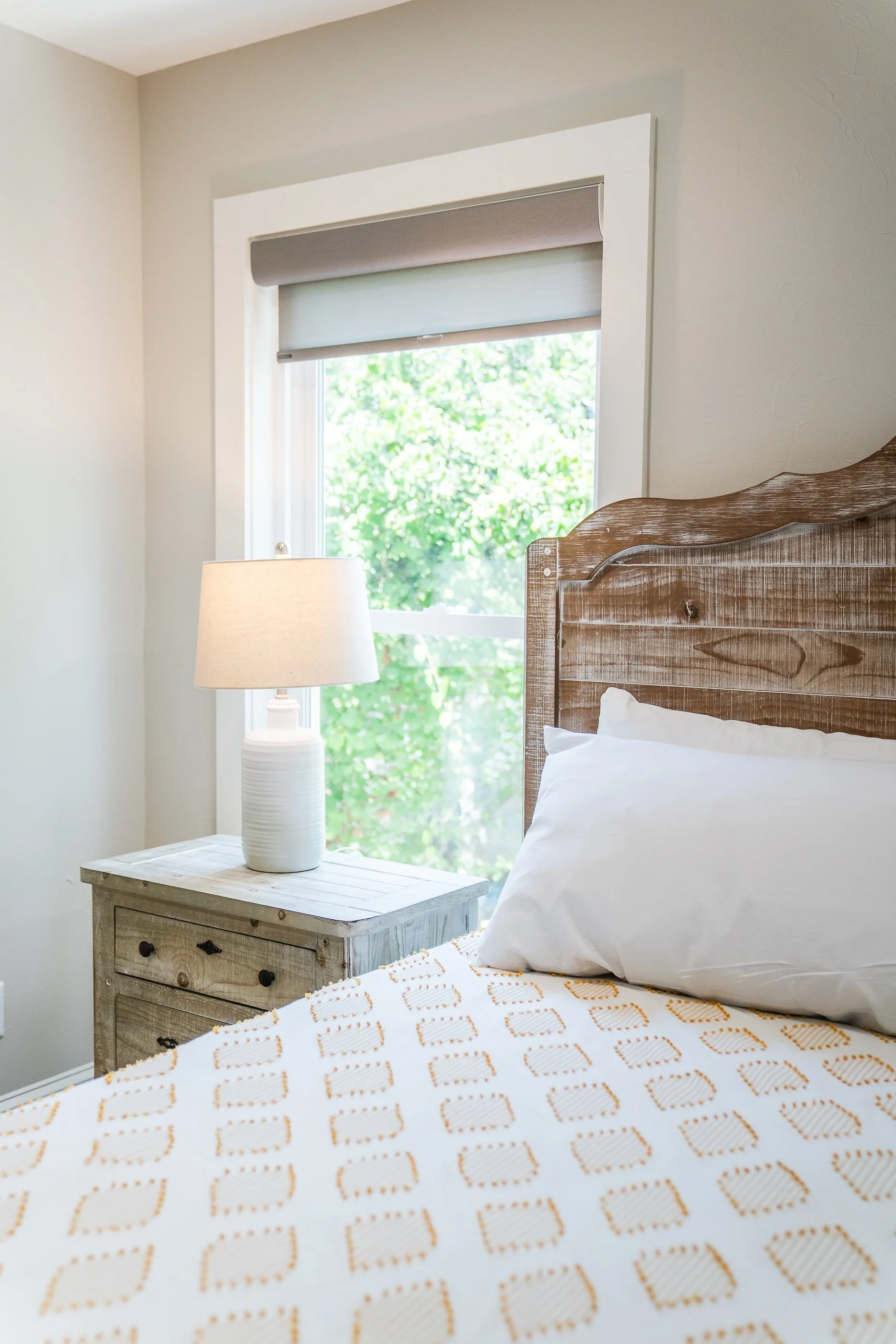 A cozy bedroom with a wooden bed, white pillow, and a patterned blanket. There is a rustic nightstand with a lamp, and a window with green foliage outside.