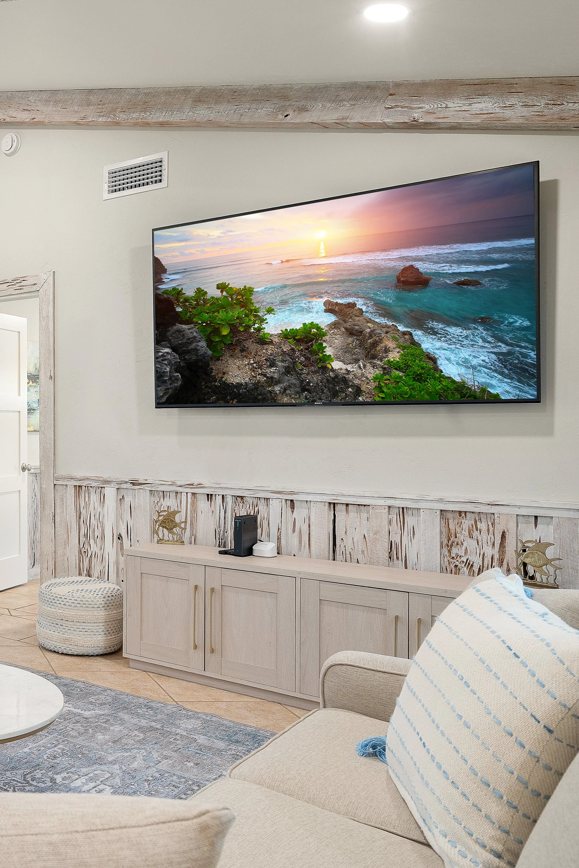 Living room with a large wall-mounted TV displaying a sunset over the ocean, beige couch with striped pillow, wooden furniture, and coastal decor.