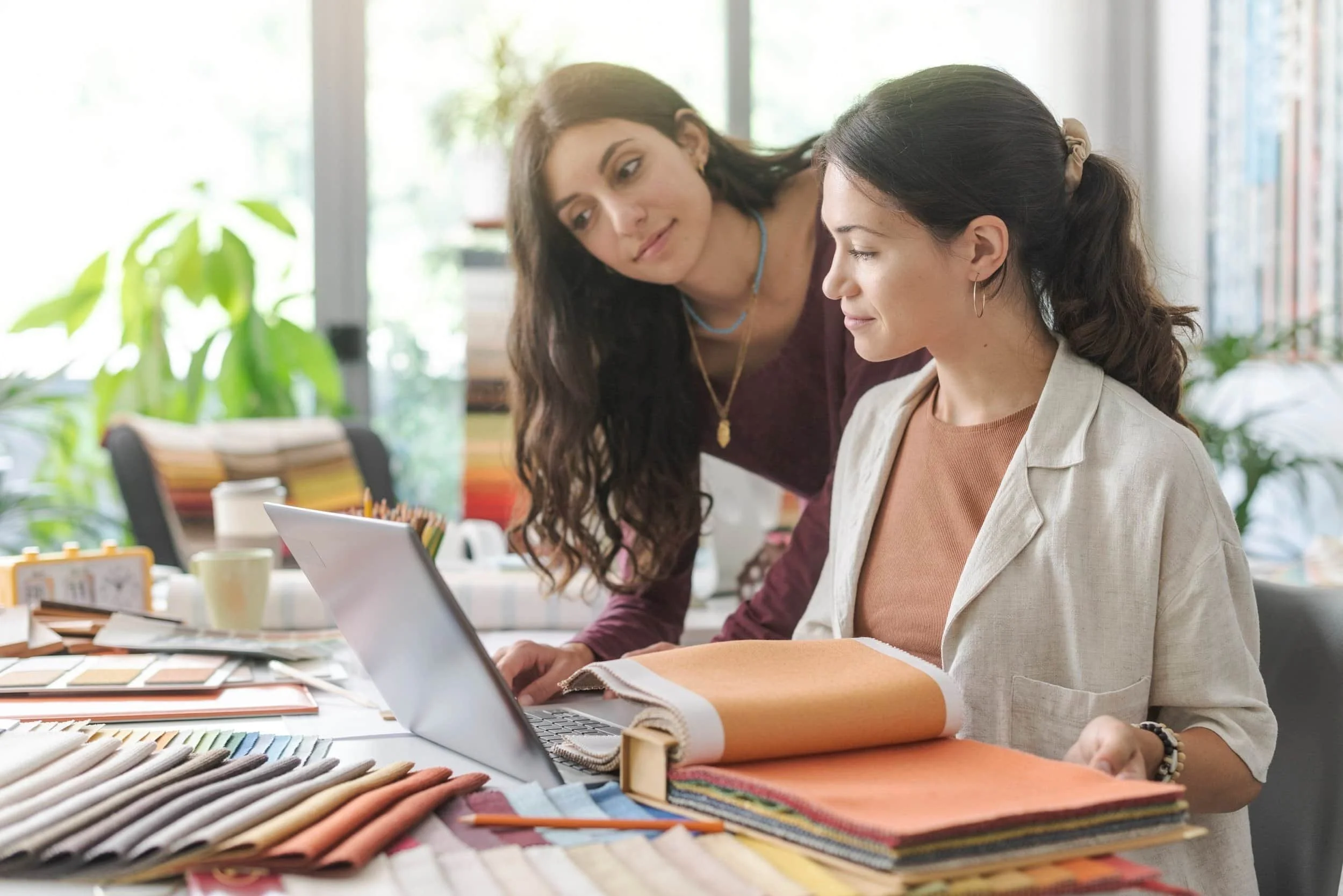 Two women working together at a desk with fabric samples, color swatches, and a laptop, in a well-lit room with plants and large windows.