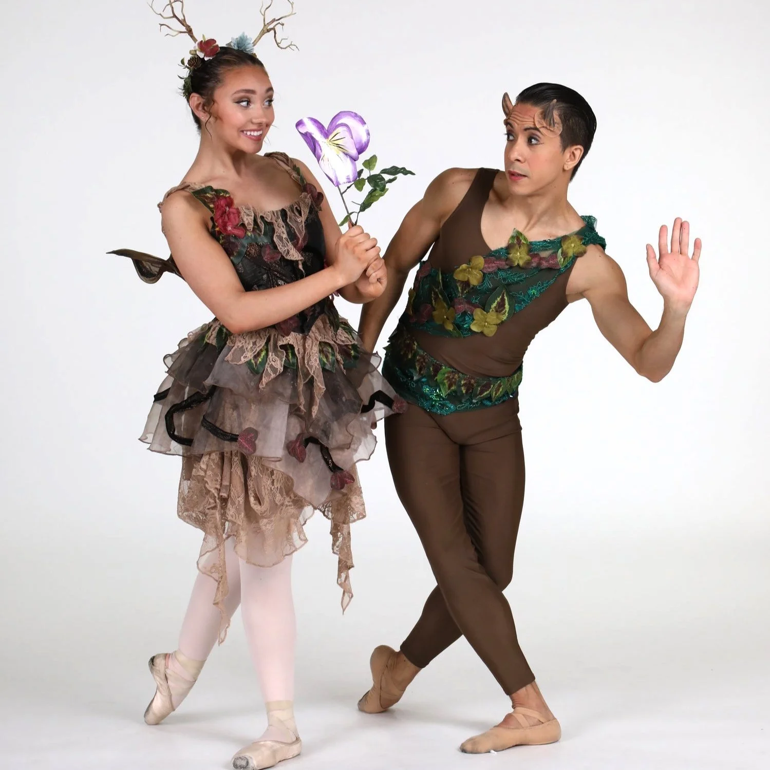 Two ballet dancers dressed as forest fairies: a woman in a floral tutu and a man in brown tights, with makeup and costumes resembling nature, pose together in a studio setting.