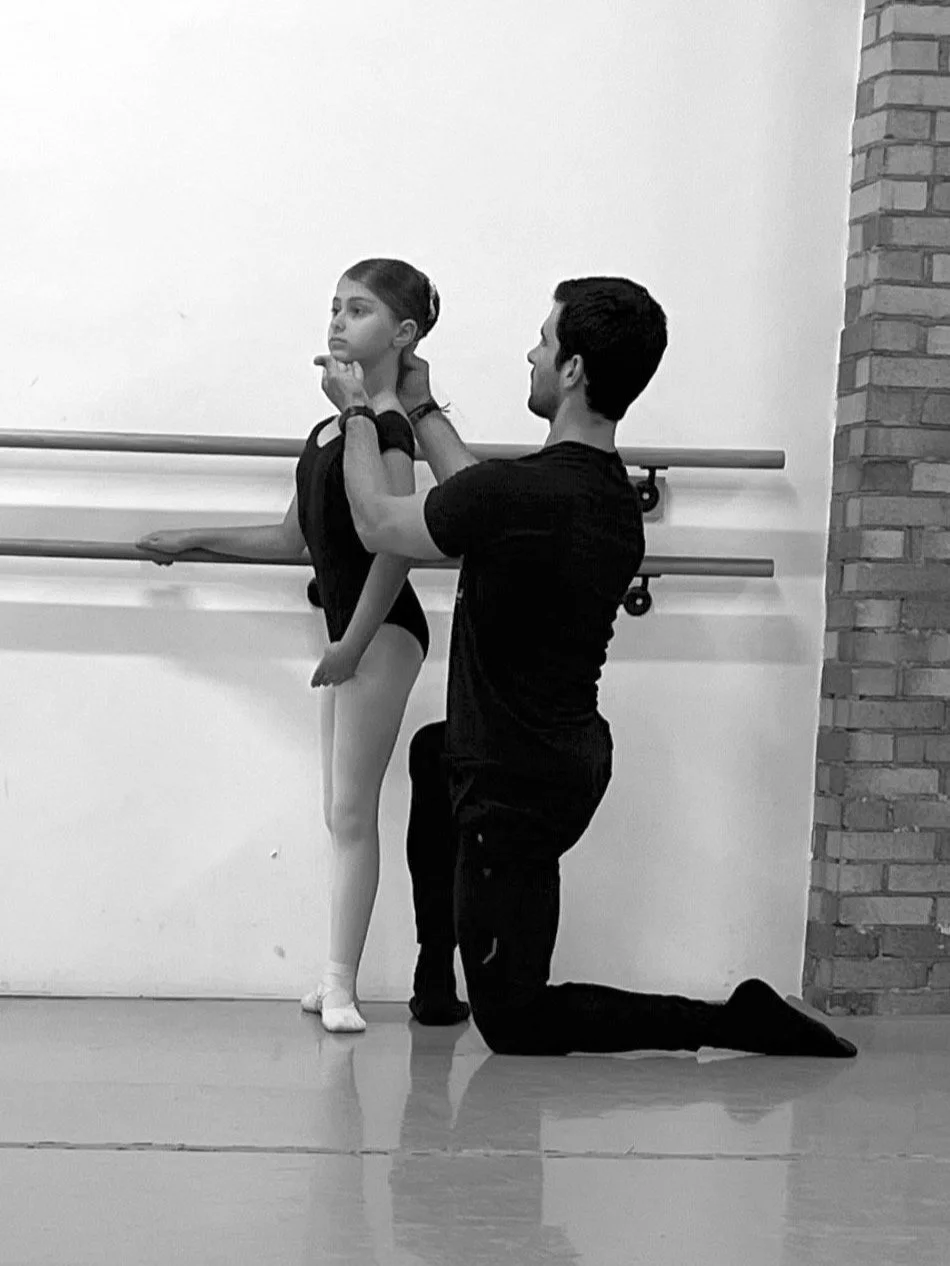 A male ballet instructor kneeling on one knee adjusting a young female ballet dancer's neck or head during a ballet class, with ballet barres and a brick wall in the background.