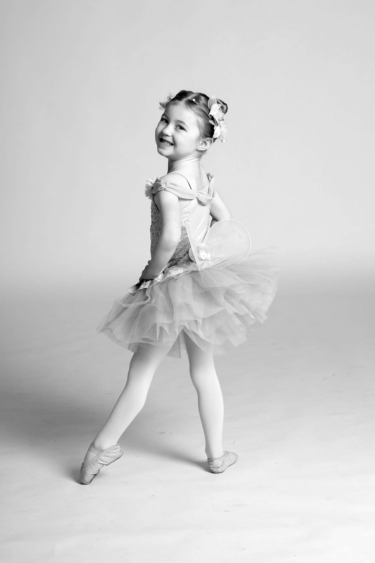 A young girl in a ballet tutu, posing en pointe with a big smile, in a studio with a plain background, in black and white.
