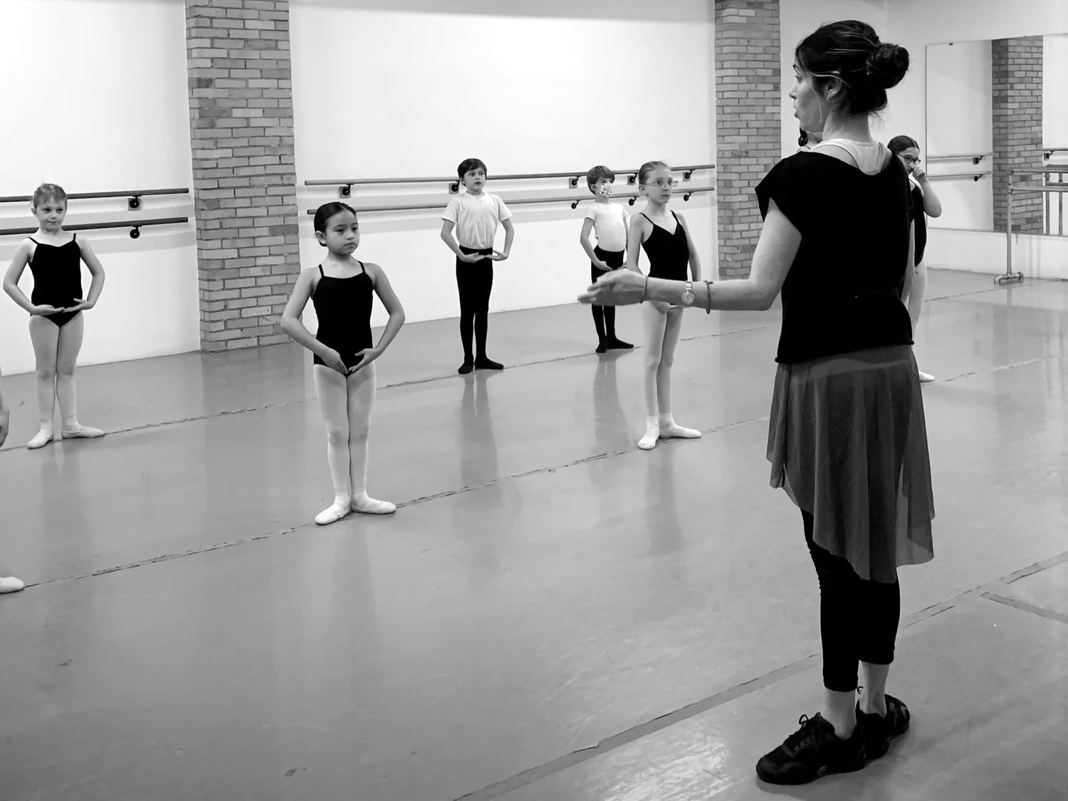 Ballet class in a dance studio with young students standing in ballet positions, listening to their instructor, a woman with her hair in a bun, wearing a black top, a skirt, and sneakers.