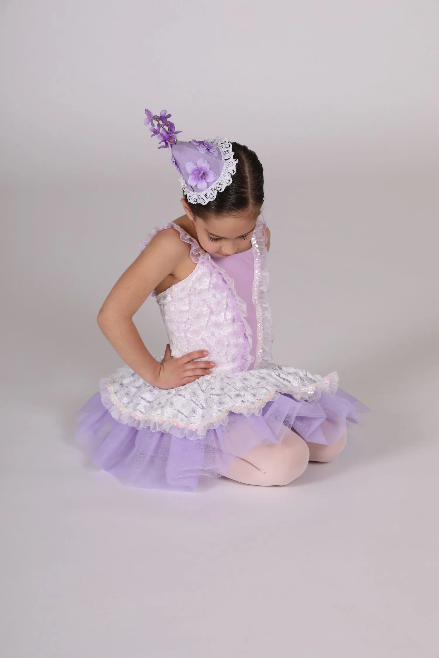 A young girl dressed in a lavender ballet costume, kneeling on the floor with her head bowed and hands on her hips, against a plain gray background.