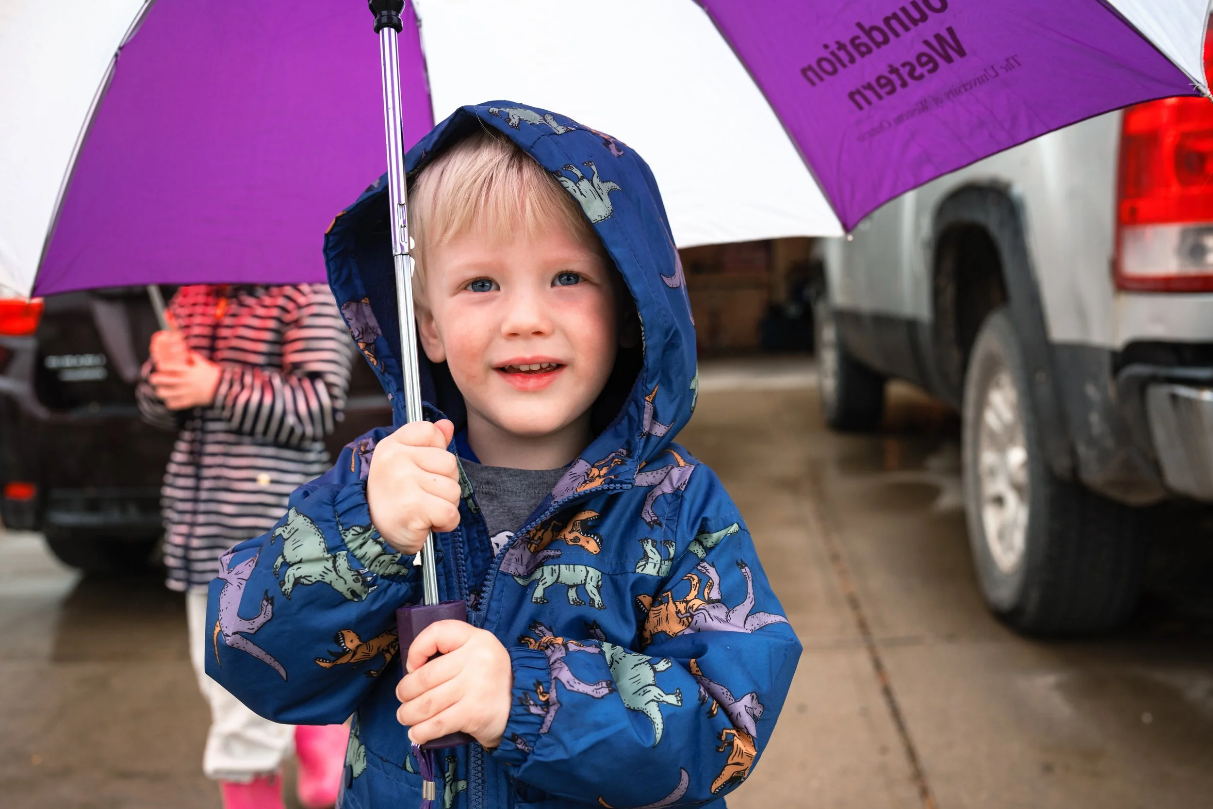 A young boy with blue eyes and blonde hair holding a purple umbrella with a blue, dinosaur-printed raincoat, standing outside in a wet driveway.