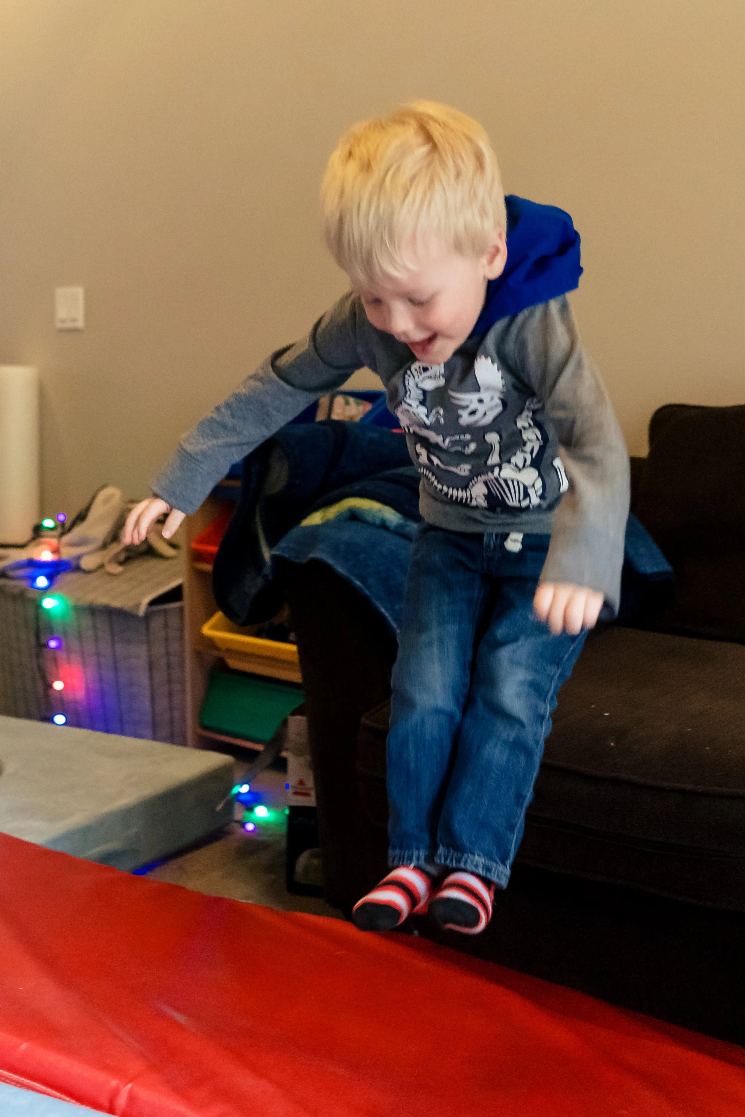 A young boy with blonde hair jumping off a couch onto a red mat, wearing a gray hoodie with a dinosaur skeleton print, jeans, and striped socks, in a living room decorated with colorful string lights.