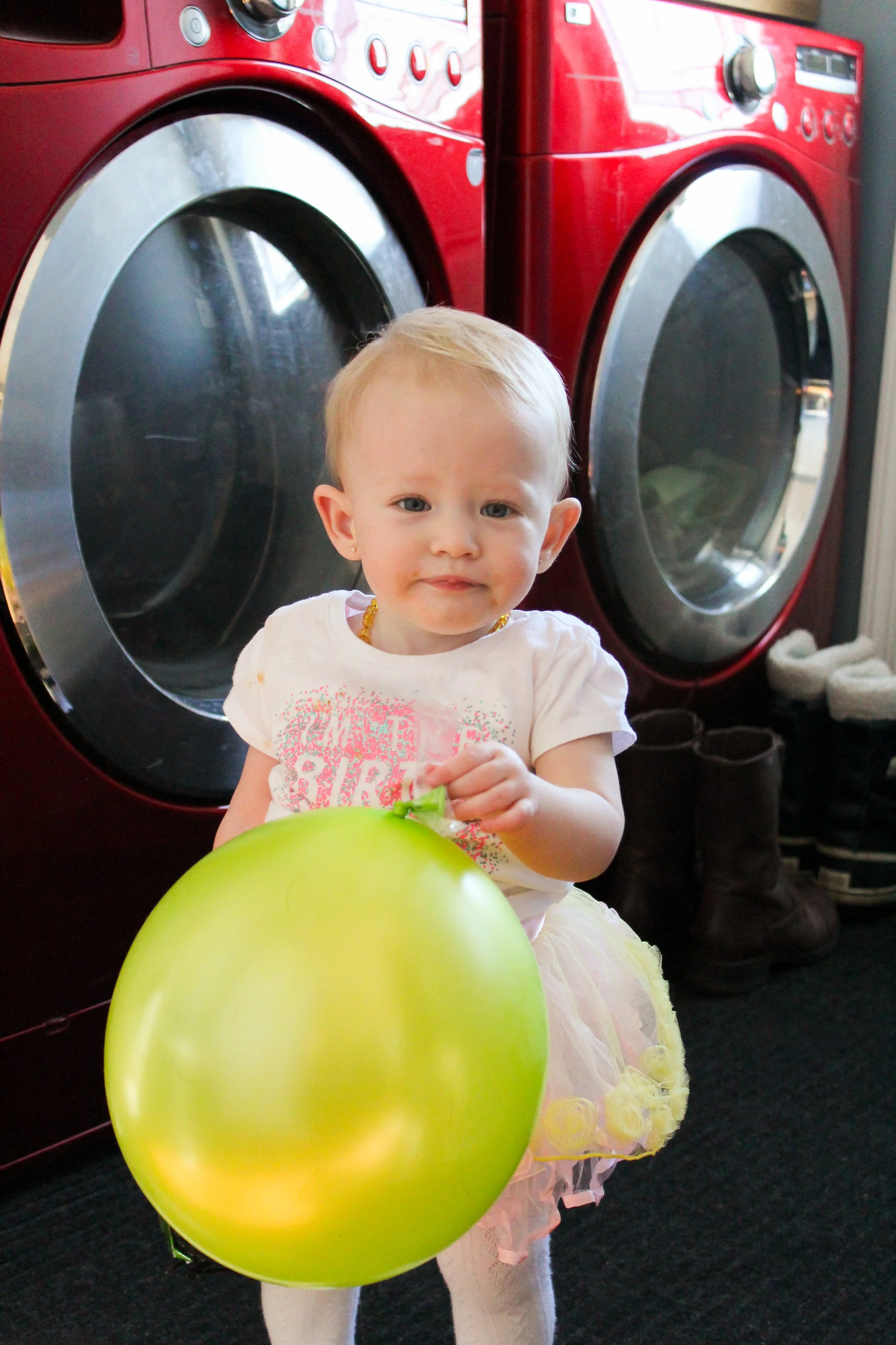 A young girl holding a green balloon in front of red washing machines in a laundry room.