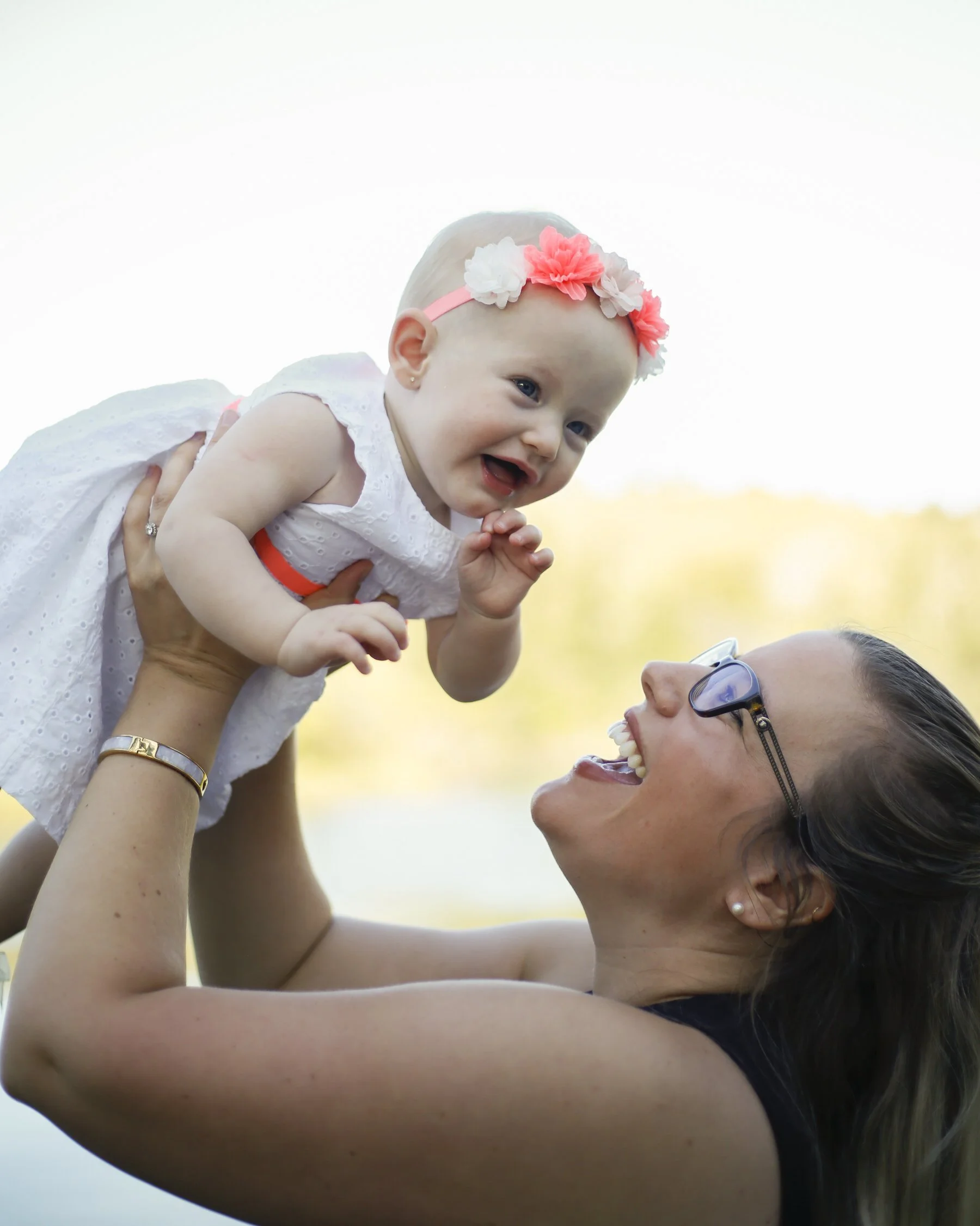 Mother lifting a smiling baby girl outdoors near a lake, with blurred trees in the background.