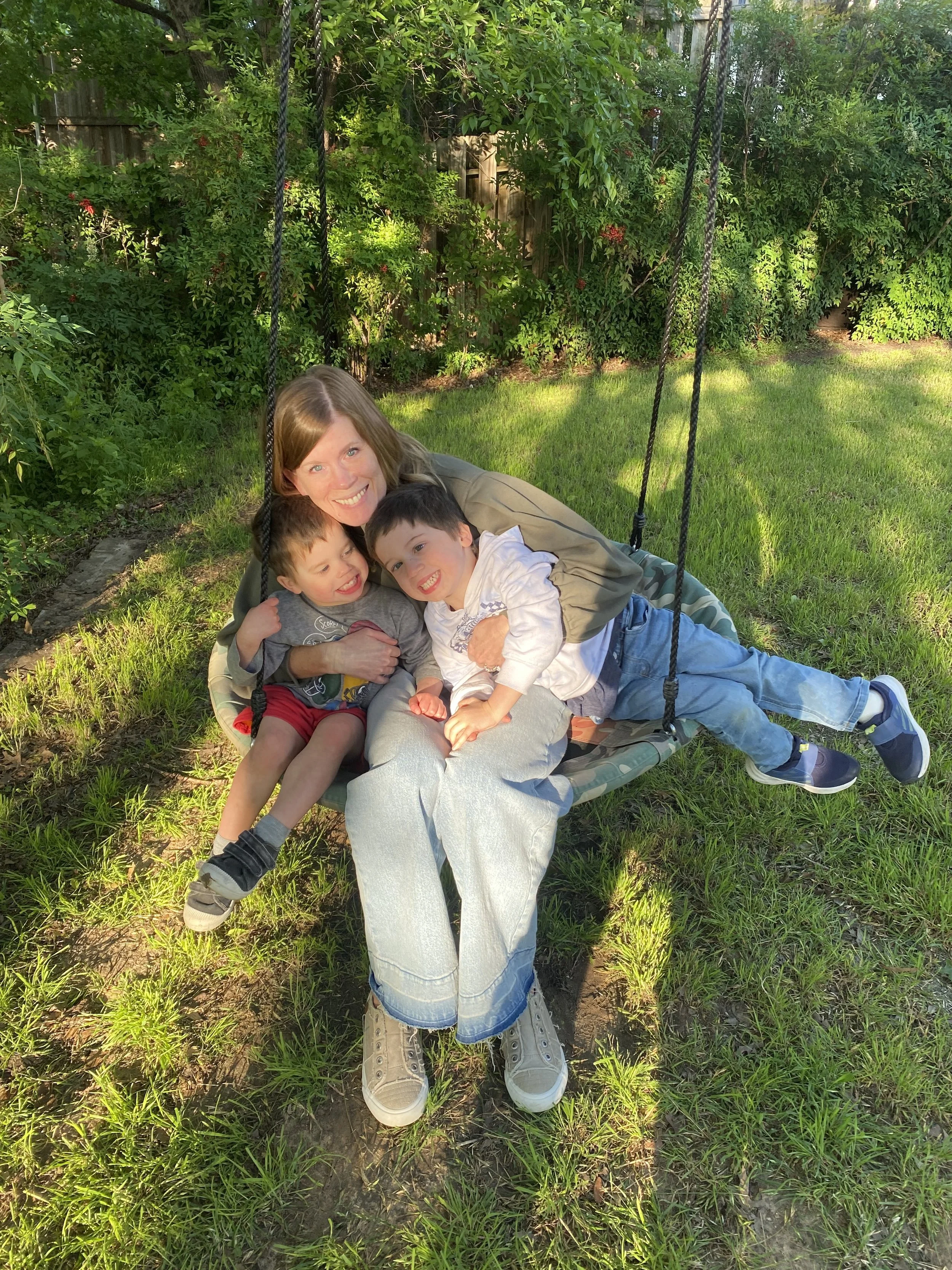 A woman and two young boys are sitting closely on a backyard porch swing, smiling and hugging each other. The setting is a sunny day with green grass and bushes in the background.