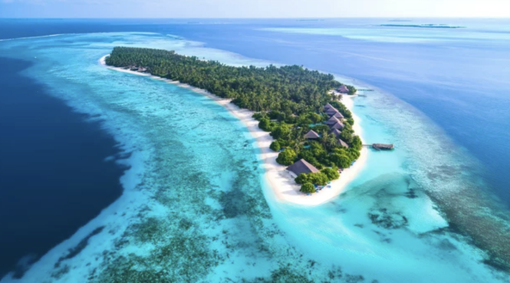 Aerial view of a tropical island with lush greenery, overwater bungalows, white sandy beaches, and surrounding turquoise ocean with coral reefs.