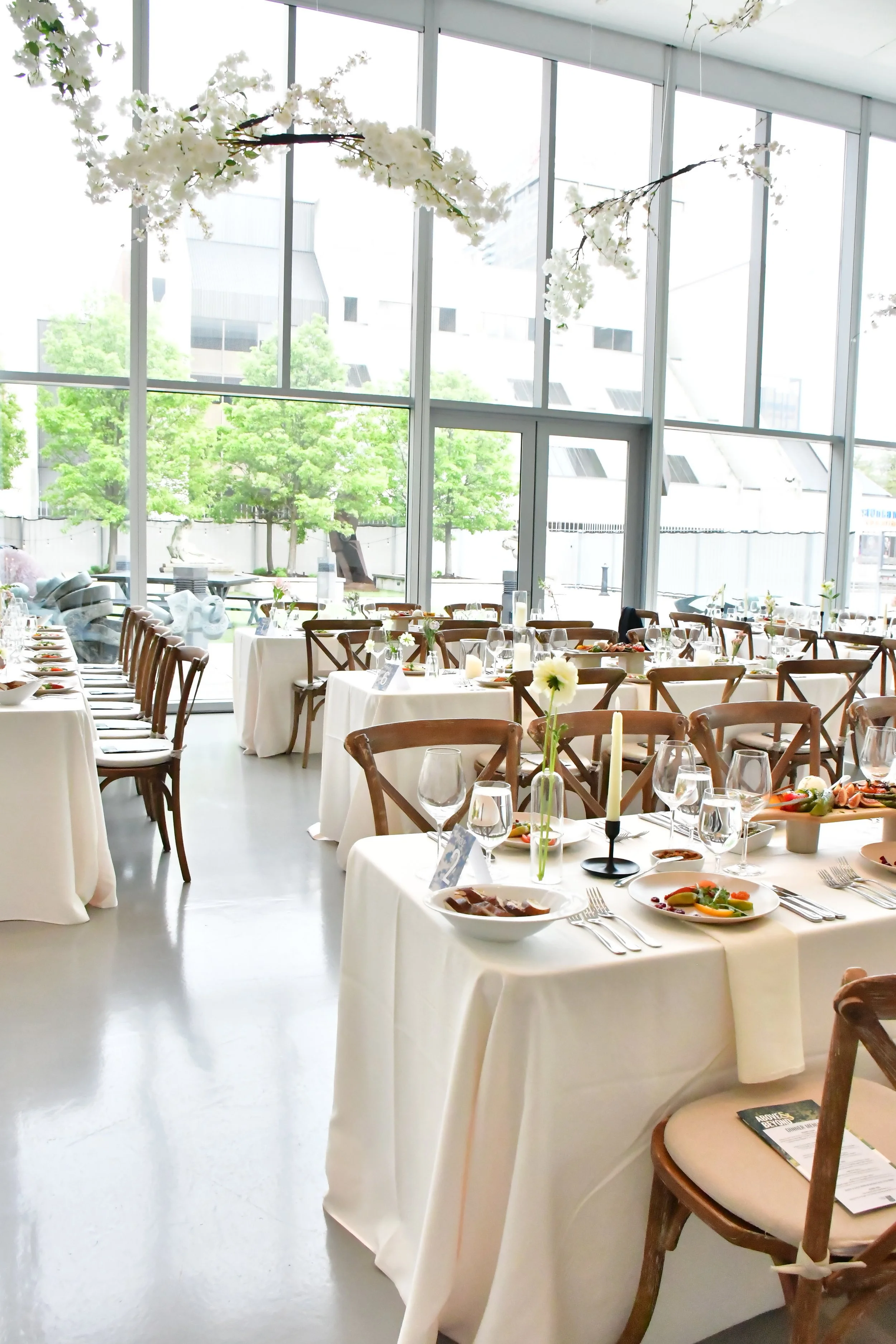 Elegant indoor event space with white-draped tables, wooden chairs, floral centerpieces, and place settings in front of large windows showing a cityscape with trees.
