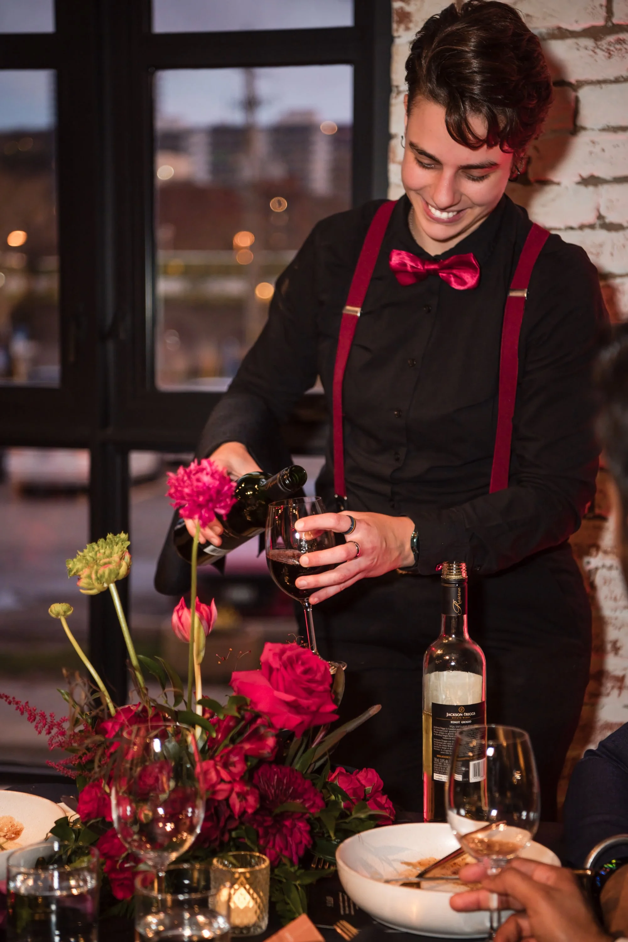 A young man with short dark hair and light skin, dressed in a black shirt with suspenders and a pink bowtie, is smiling as he pours wine into a glass. The scene appears to be a dinner or celebration in a restaurant or a decorated dining area, with a 
