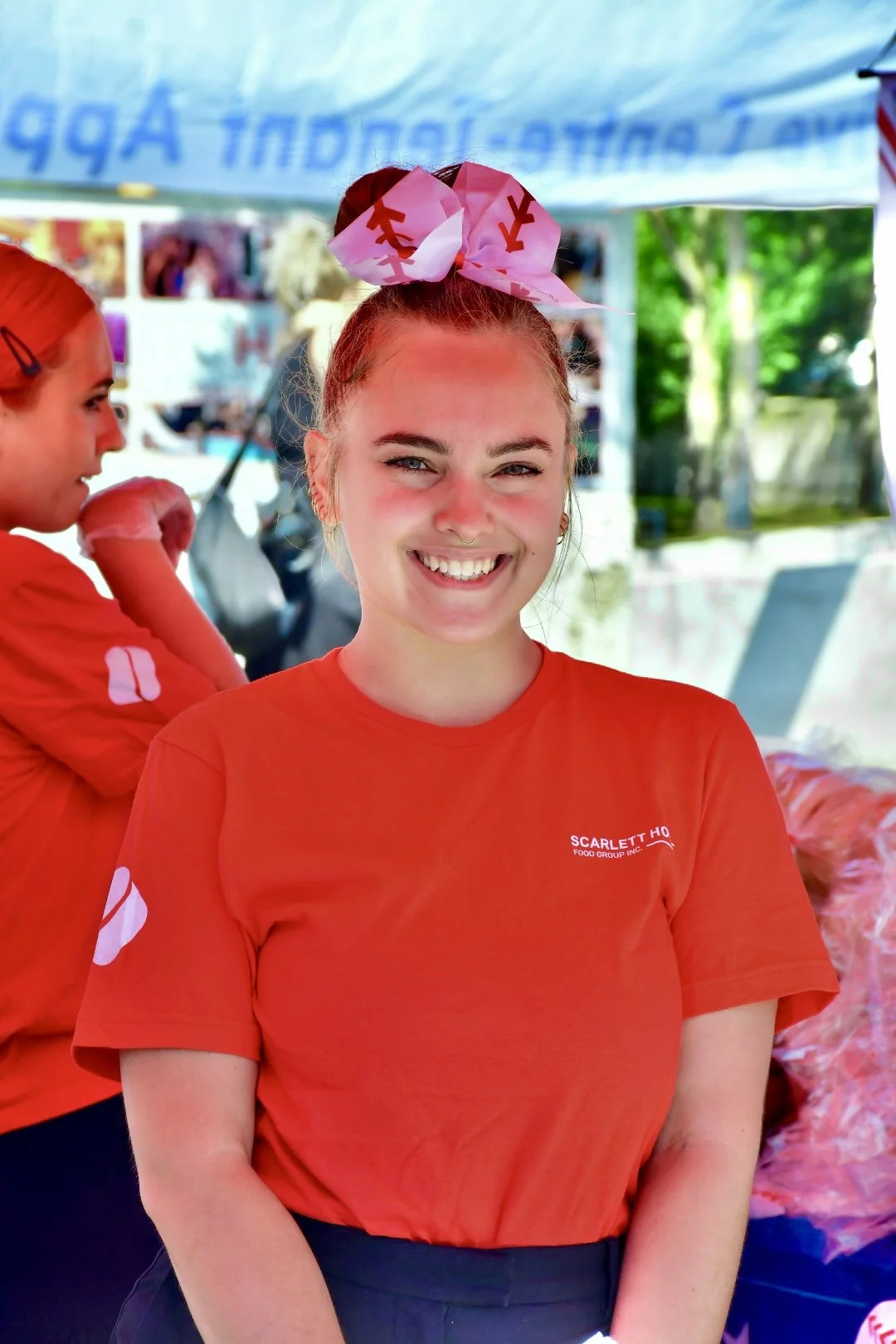A young woman smiling, wearing a red t-shirt with the Scarlett logo printed on it and a baseball themed bow in her hair, standing outdoors at a booth with food, with another person in red shirt in the background.