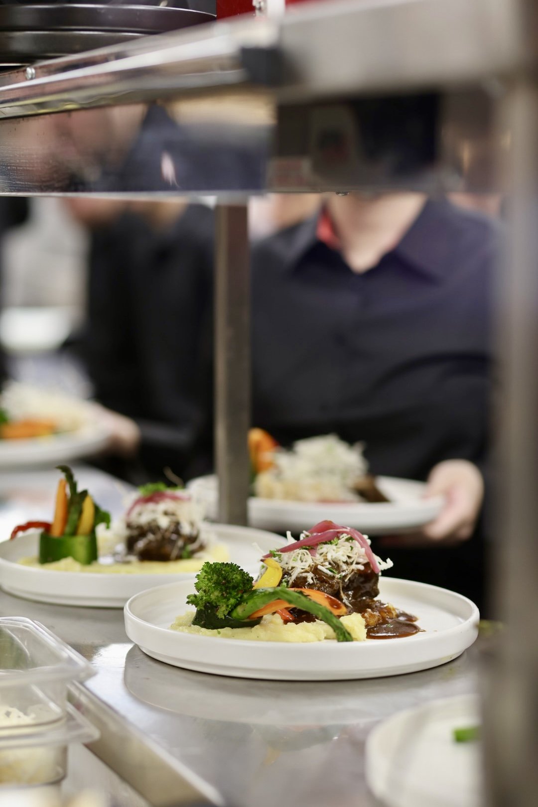 Plated entrees on a serving line in a catering kitchen with a server in a black uniform in the background.