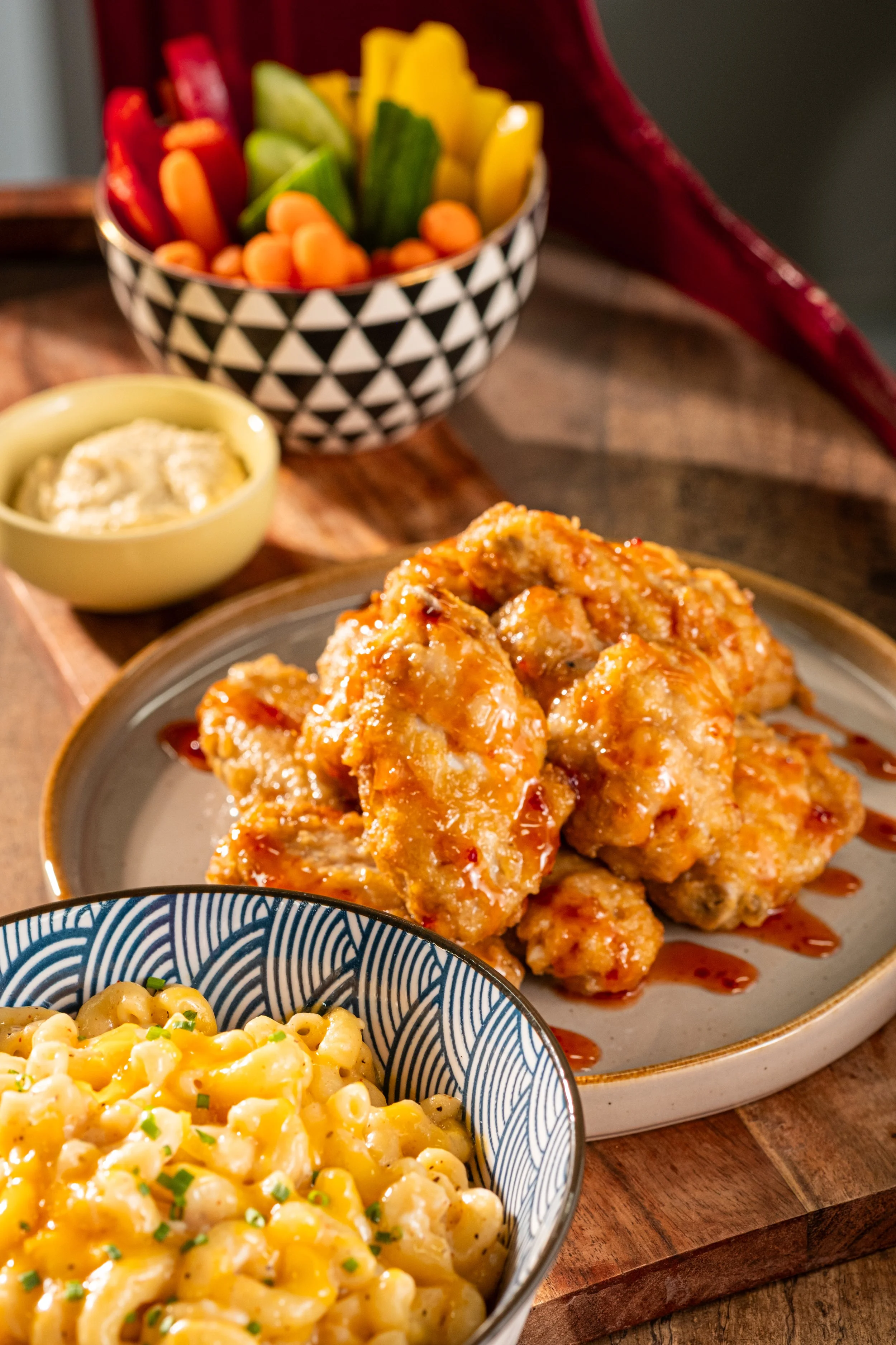 Fried chicken wings with sauce, macaroni and cheese, vegetable platter with sliced peppers and cucumbers, and a small bowl of dipping sauce on a wooden table.