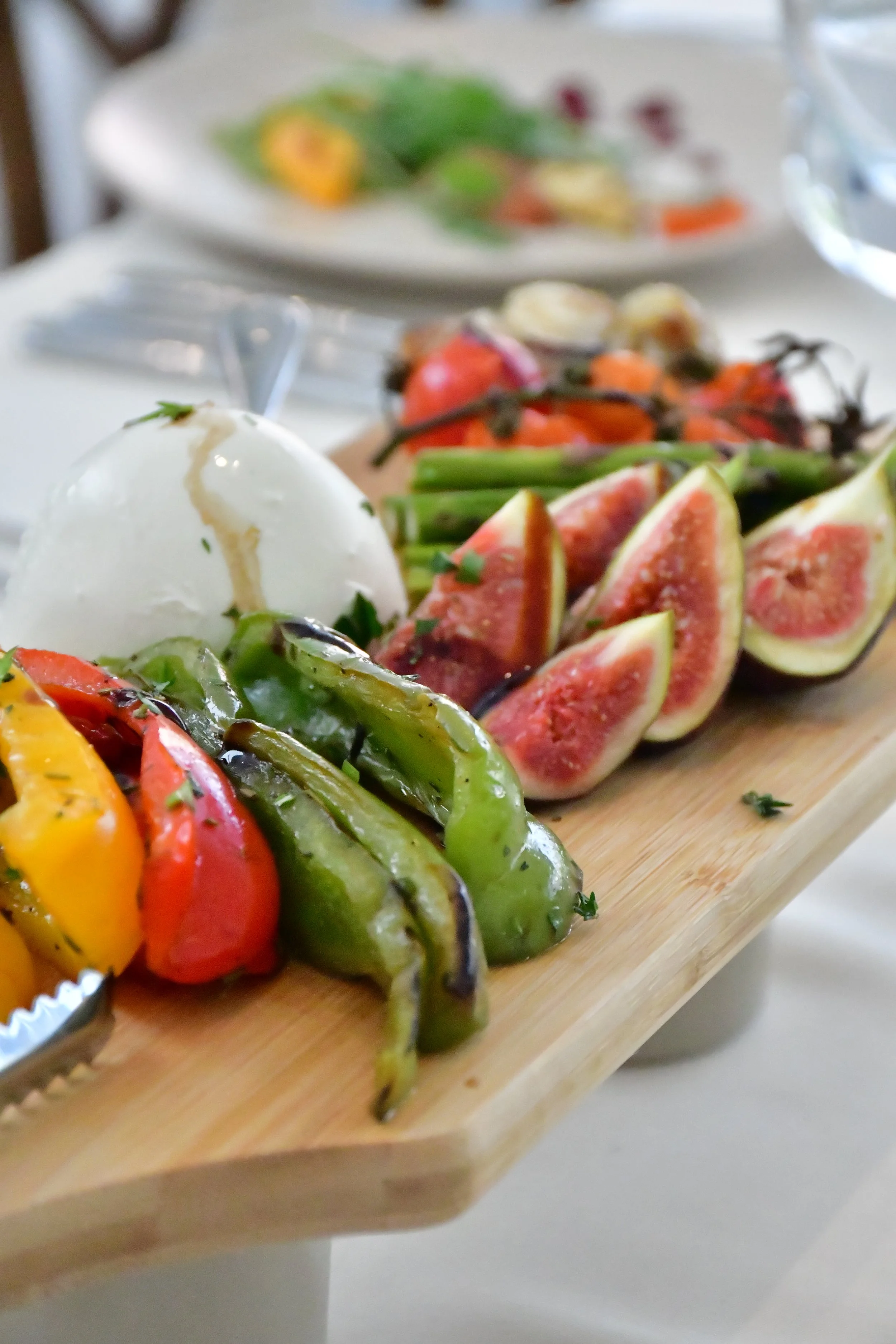 A wooden serving board with grilled colorful bell peppers, fresh figs, and a ball of burrata cheese with a drizzle of balsamic reduction, with a blurred plate of salad in the background.