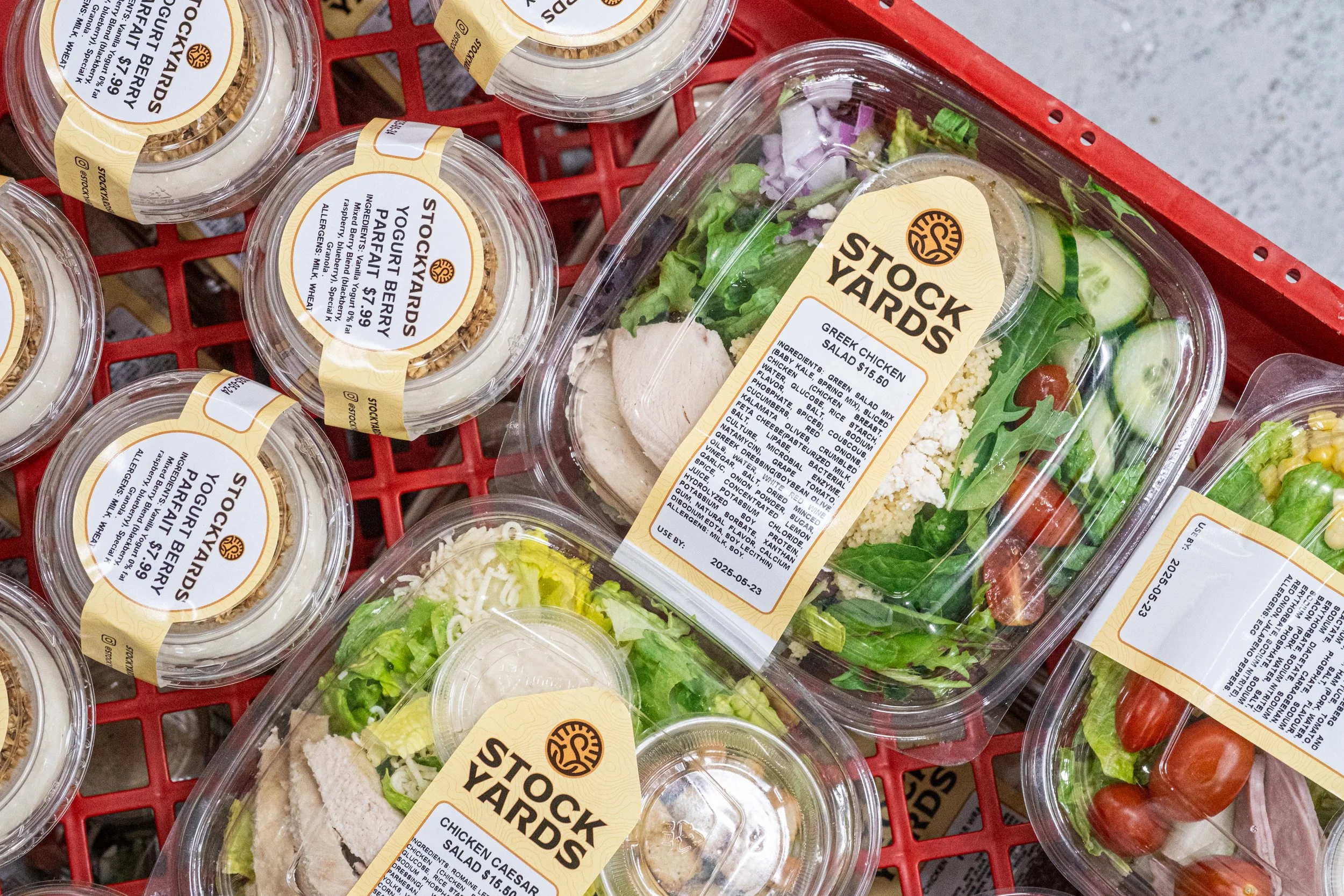 Pre-packaged salads and snack containers on a red rack, featuring Chicken Caesar Salad and yogurt parfaits prepared for the Stock Yards cafe at the Waterloo International Airport.
