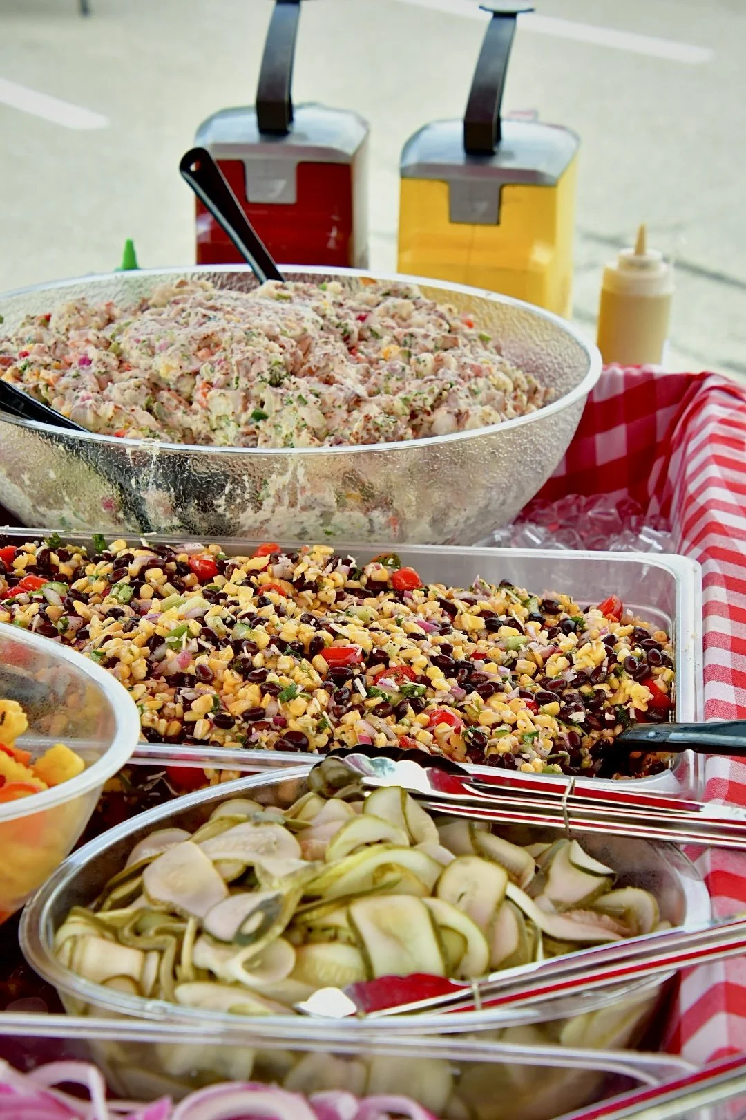 A variety of salads and garnishes in clear bowls on a table with red checkered cloth at an outdoor food station.