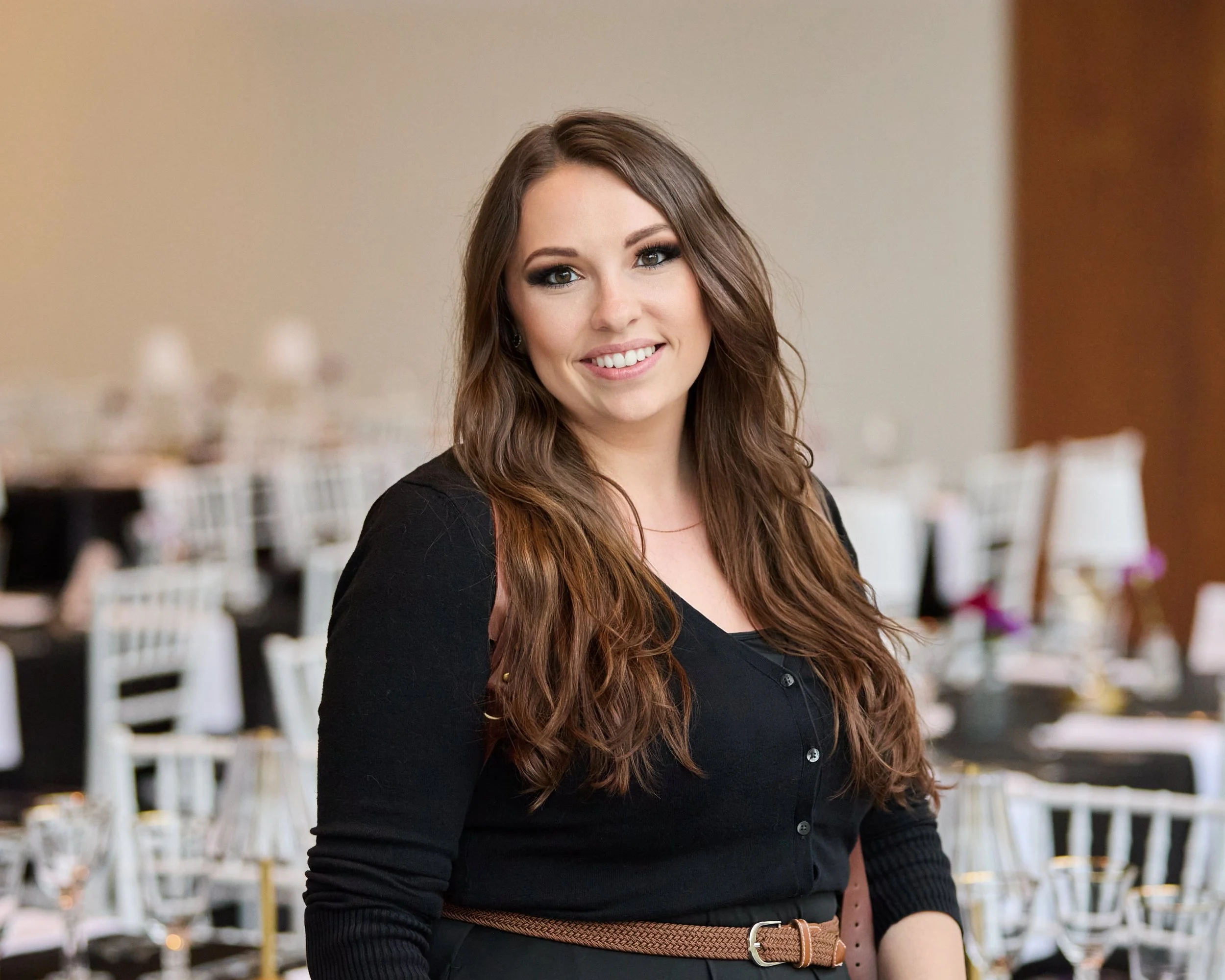 A portrait of a young woman with long, wavy brown hair with white chivari chairs and tables set for a formal event in the background. This is Deanna, the catering & events marketing administrator.