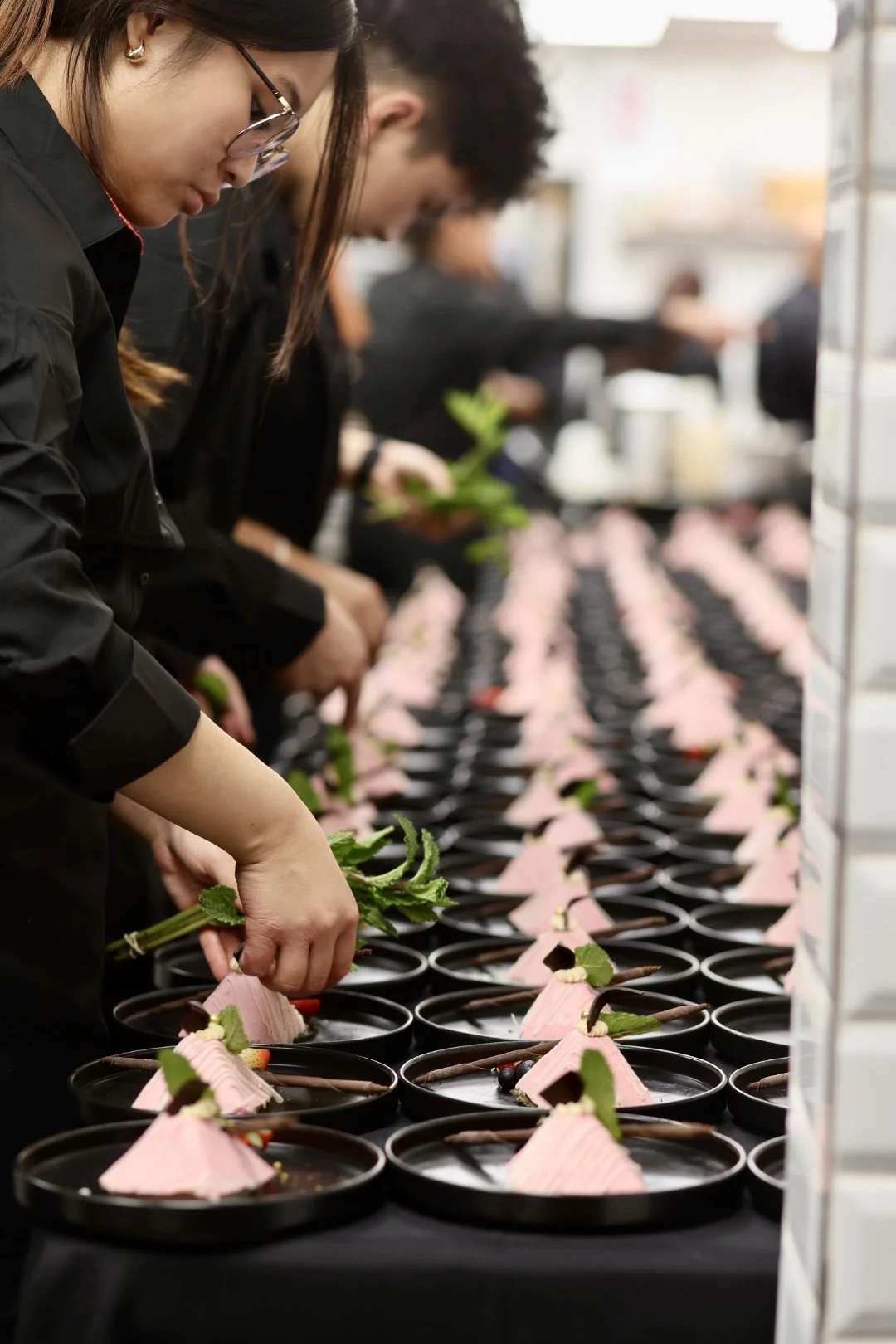 Scarlett catering staff in a catering kitchen plating desserts arranged in a row, with pink mounds, chocolate decorations, and green garnishes, at a special event.