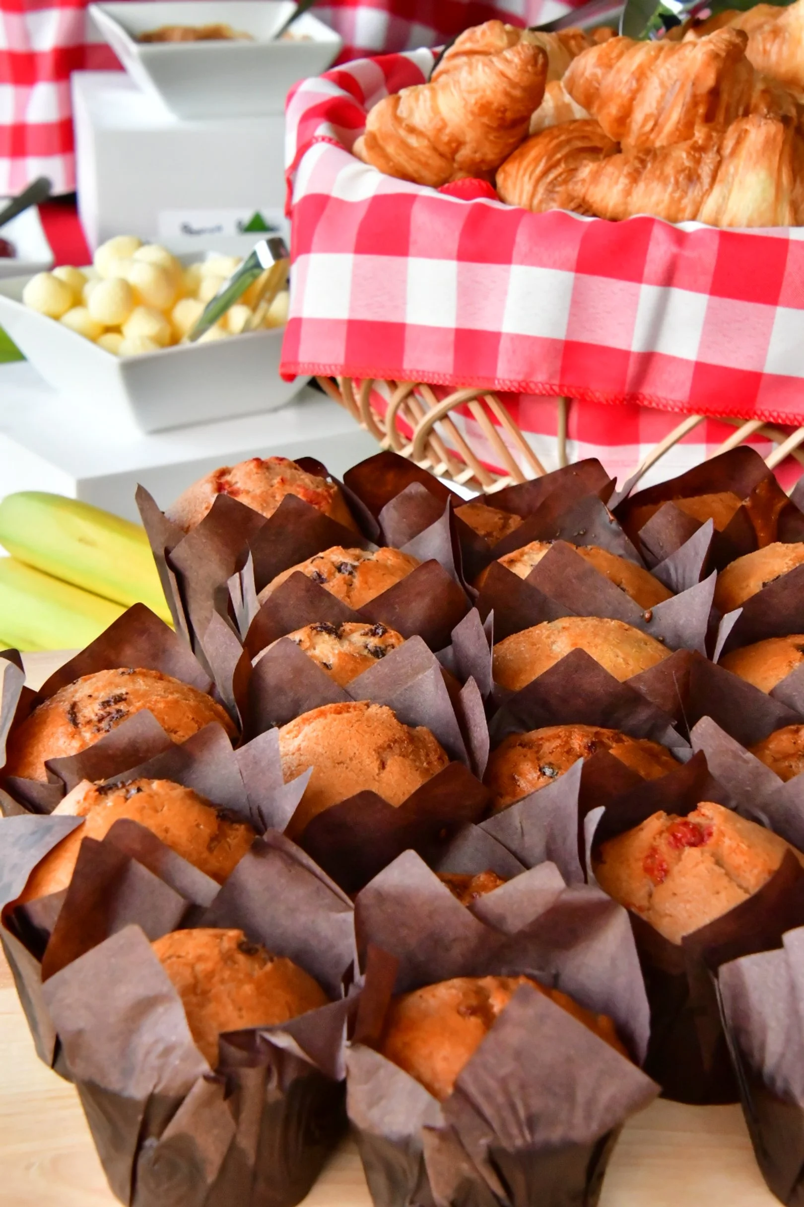 A tray of muffins in paper liners, a basket of croissants with a red and white checkered cloth, bananas, and other breakfast items on a table.
