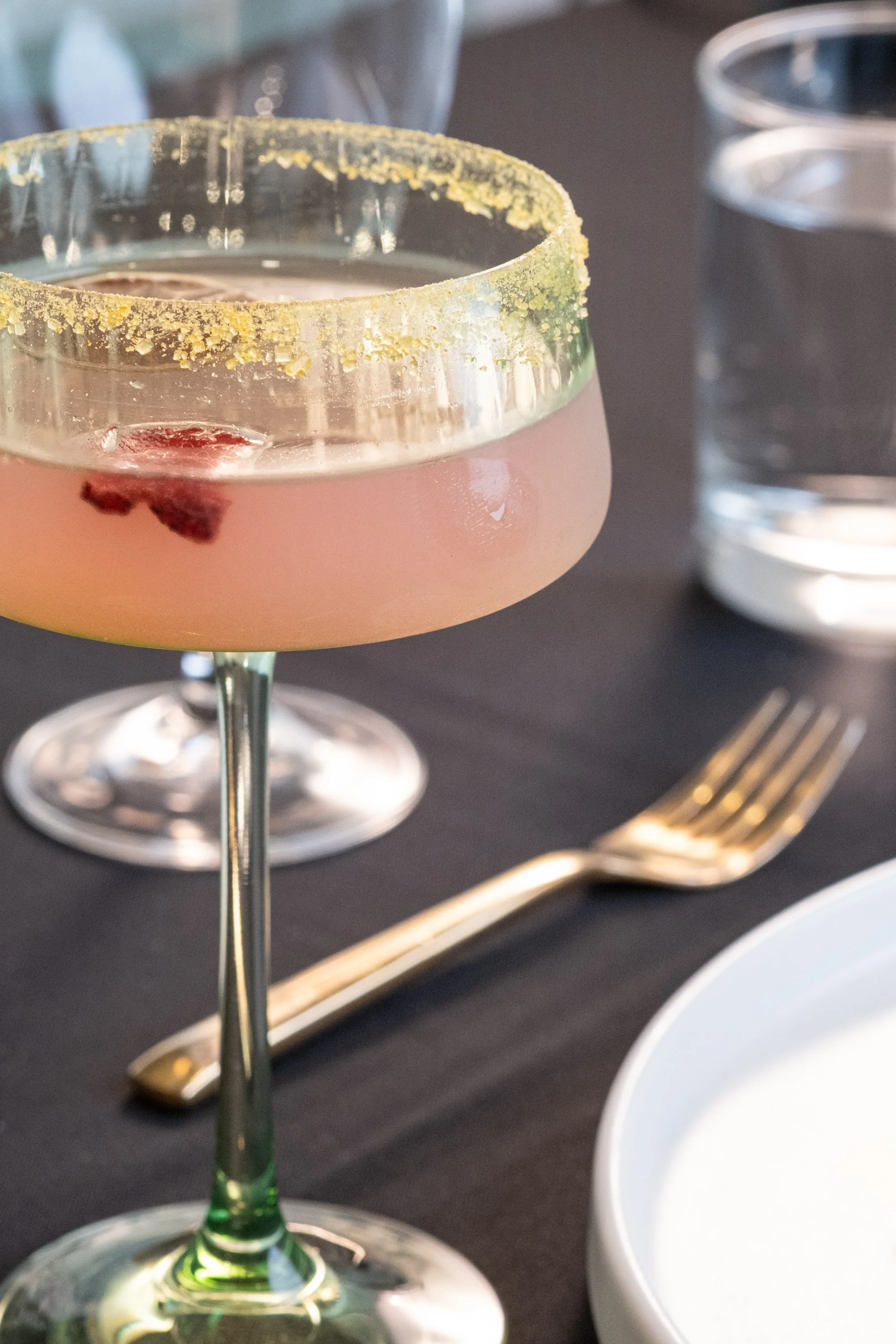 A pink cocktail in a margarita glass with a salted rim, garnished with a small fruit piece, on a black table with a fork, knife, and a white ceramic dish nearby.