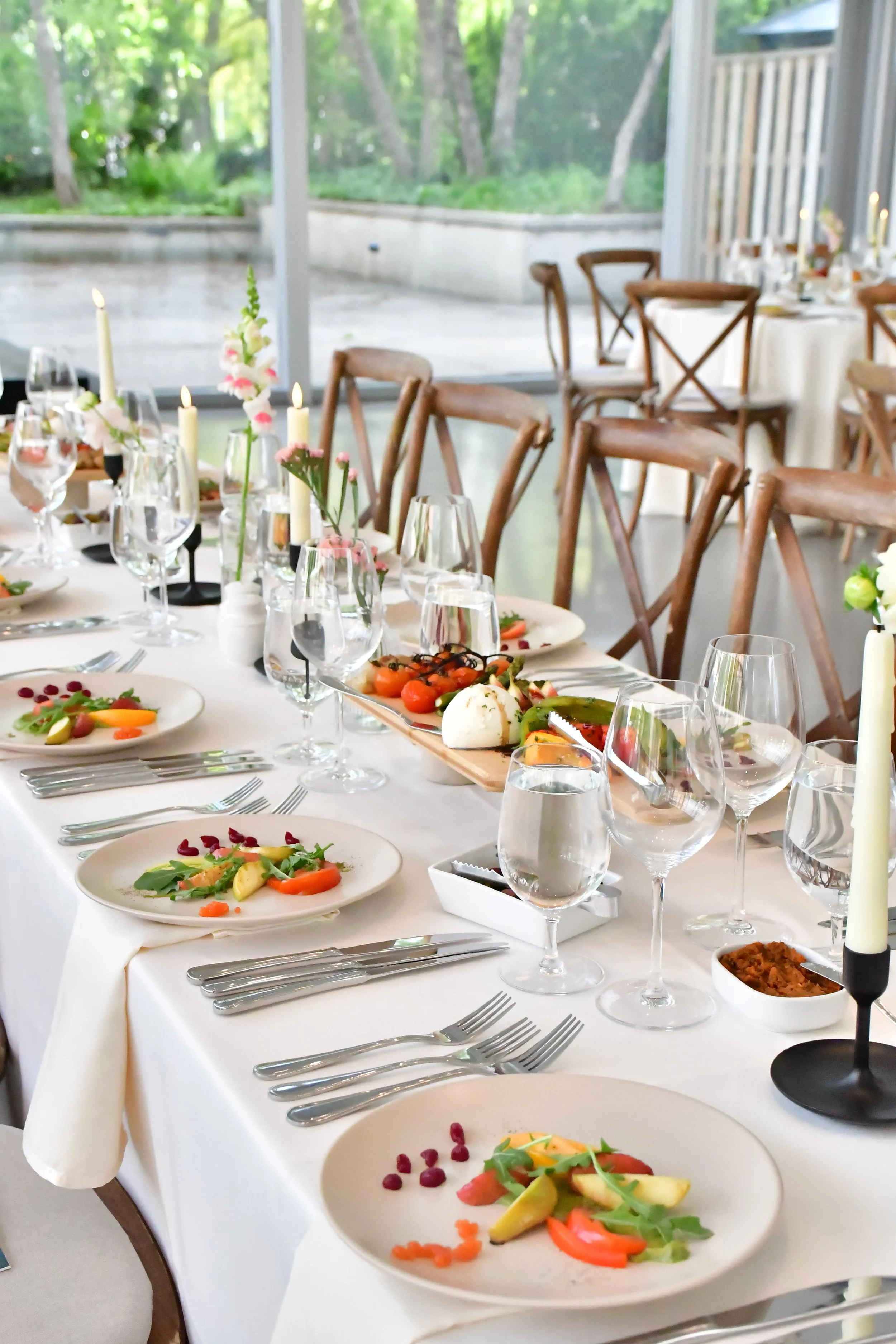 Elegant dining table set with plates of salad, wine glasses, candles, and a wooden interior with greenery outside.