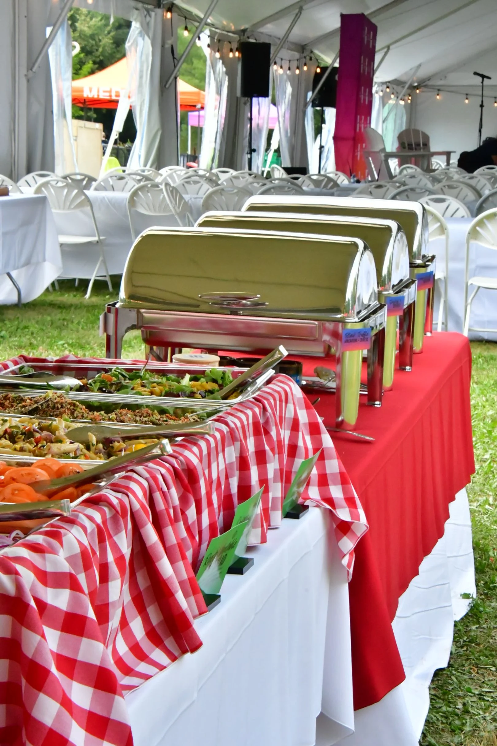 A buffet table with stainless steel chafing dishes and various salads in bowls, set up outdoors under a large white tent with tables and chairs. Red and white checkered cloths drape the table, and there are string lights hanging overhead.