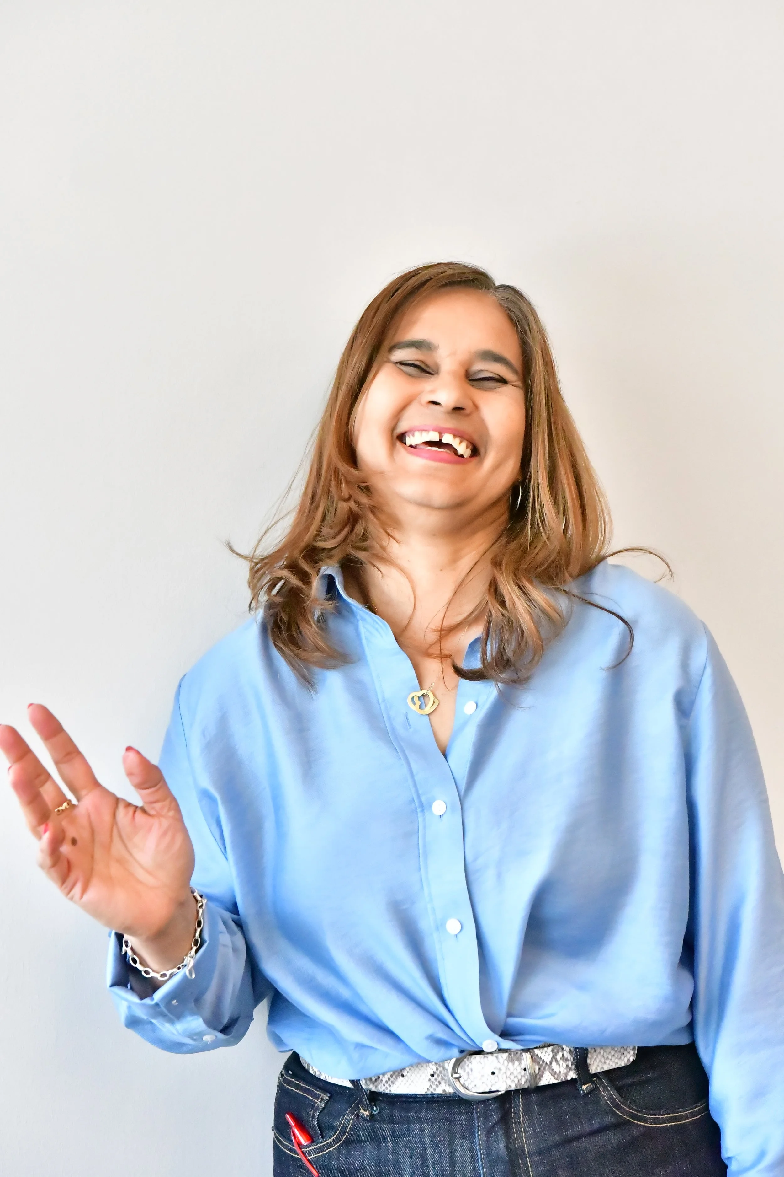 A woman smiling and waving, wearing a light blue button-up shirt, dark jeans, and a white belt, standing against a plain white wall.