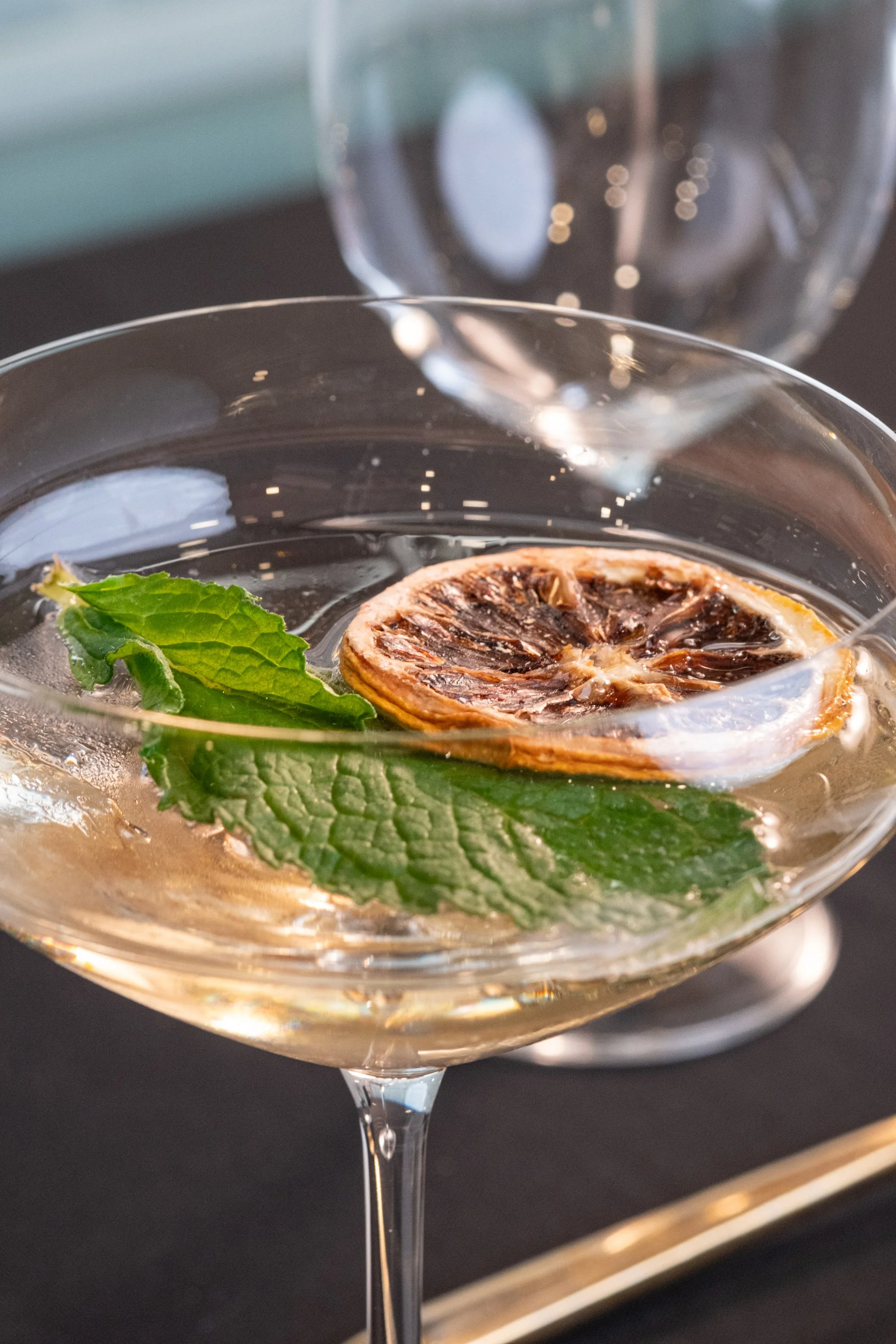 Close-up of a cocktail glass with a lemon slice and mint leaves, with an empty glass in the background.