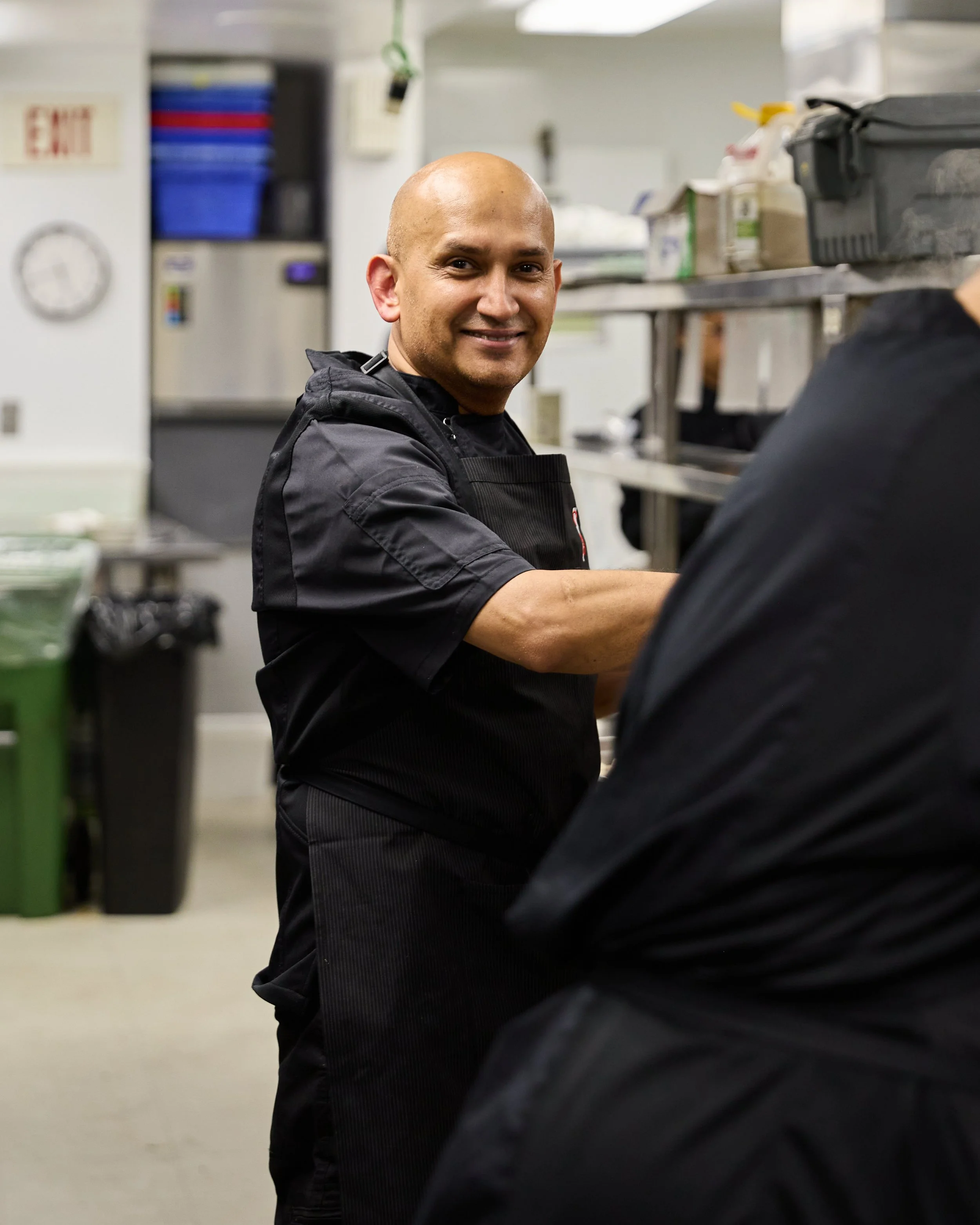 A male chef with a bald head and a slight smile, wearing a black uniform, in a commercial kitchen.