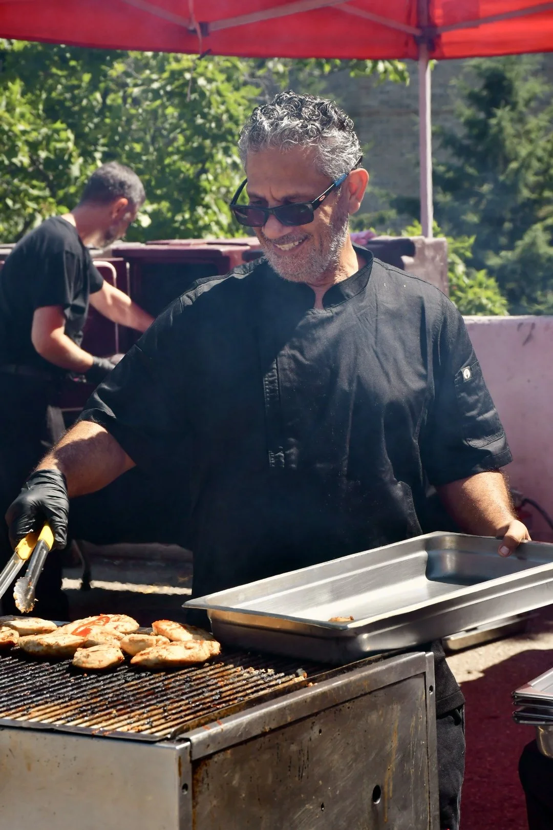 A smiling male chef wearing black sunglasses and a black chef's uniform grilling chicken breasts outdoors under a red canopy, with another person in the background also cooking.