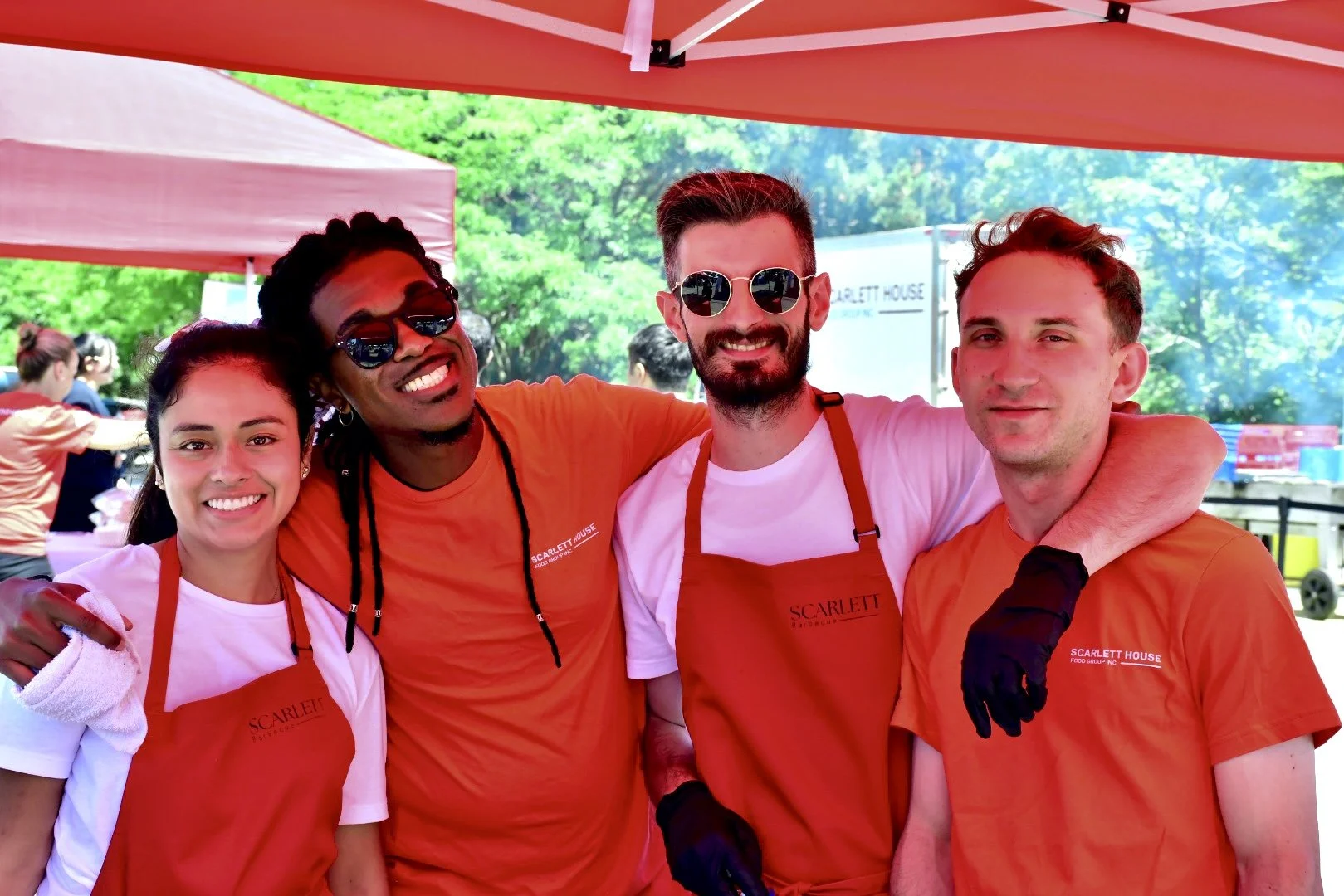 Four staff members smiling and with arms around each other at an outdoor event, wearing red aprons with 'Scarlett' logo, under red canopy, with green trees in the background.