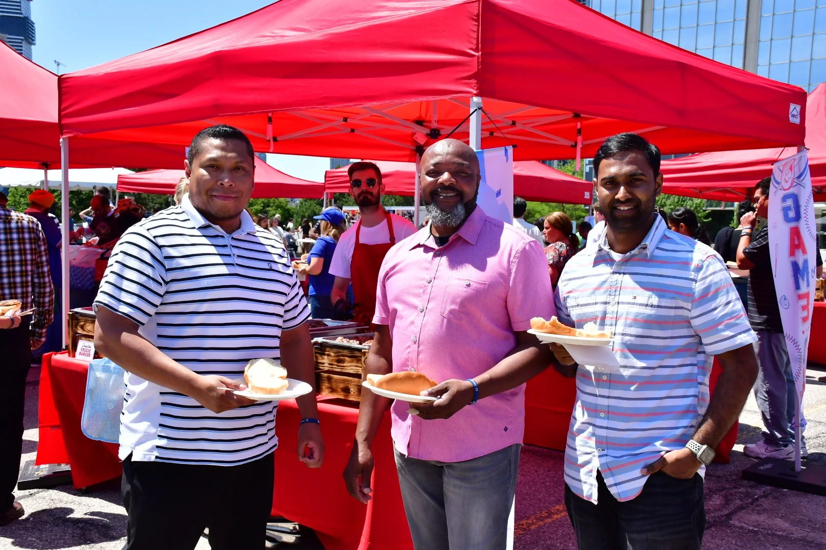 Three men holding plates of food at an outdoor event with red tents and other attendees in the background.