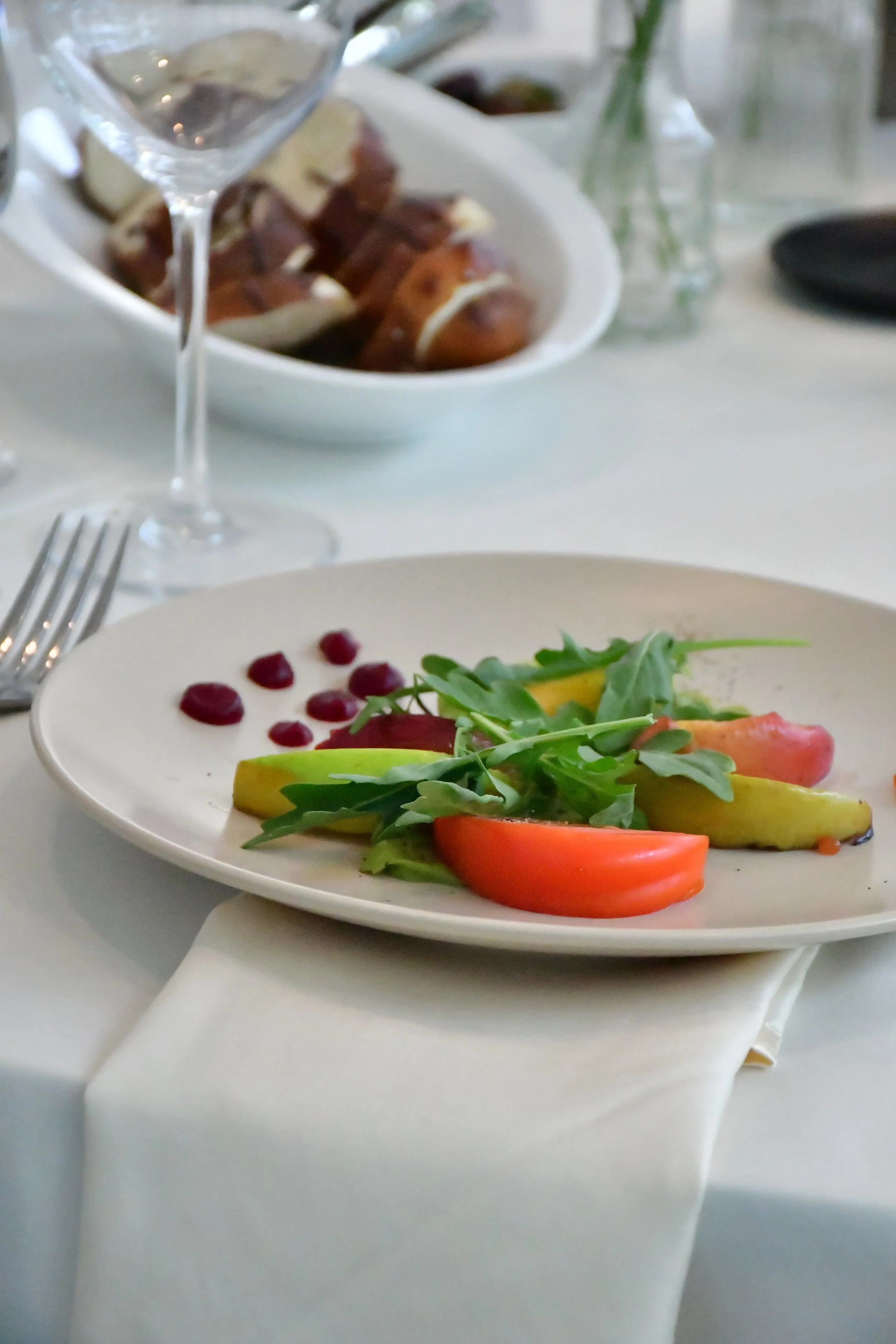 A white plate with a small salad of arugula, sliced heirloom tomatoes, and marinated pears, garnished with a few drops of a red sauce, on a white tablecloth. 