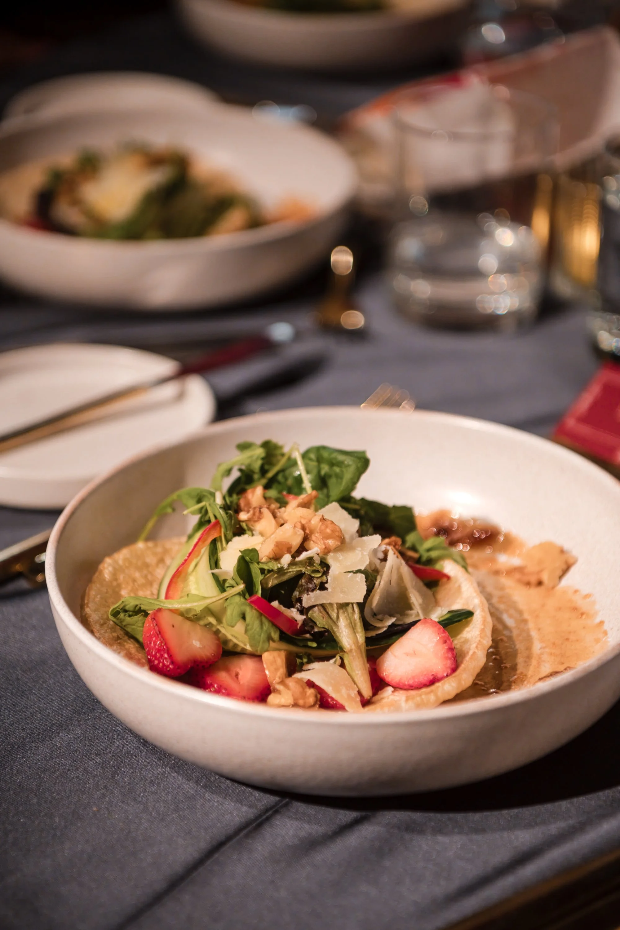 A bowl of salad with strawberries, greens, and shaved cheese on top, placed on a dark table with a fork nearby.