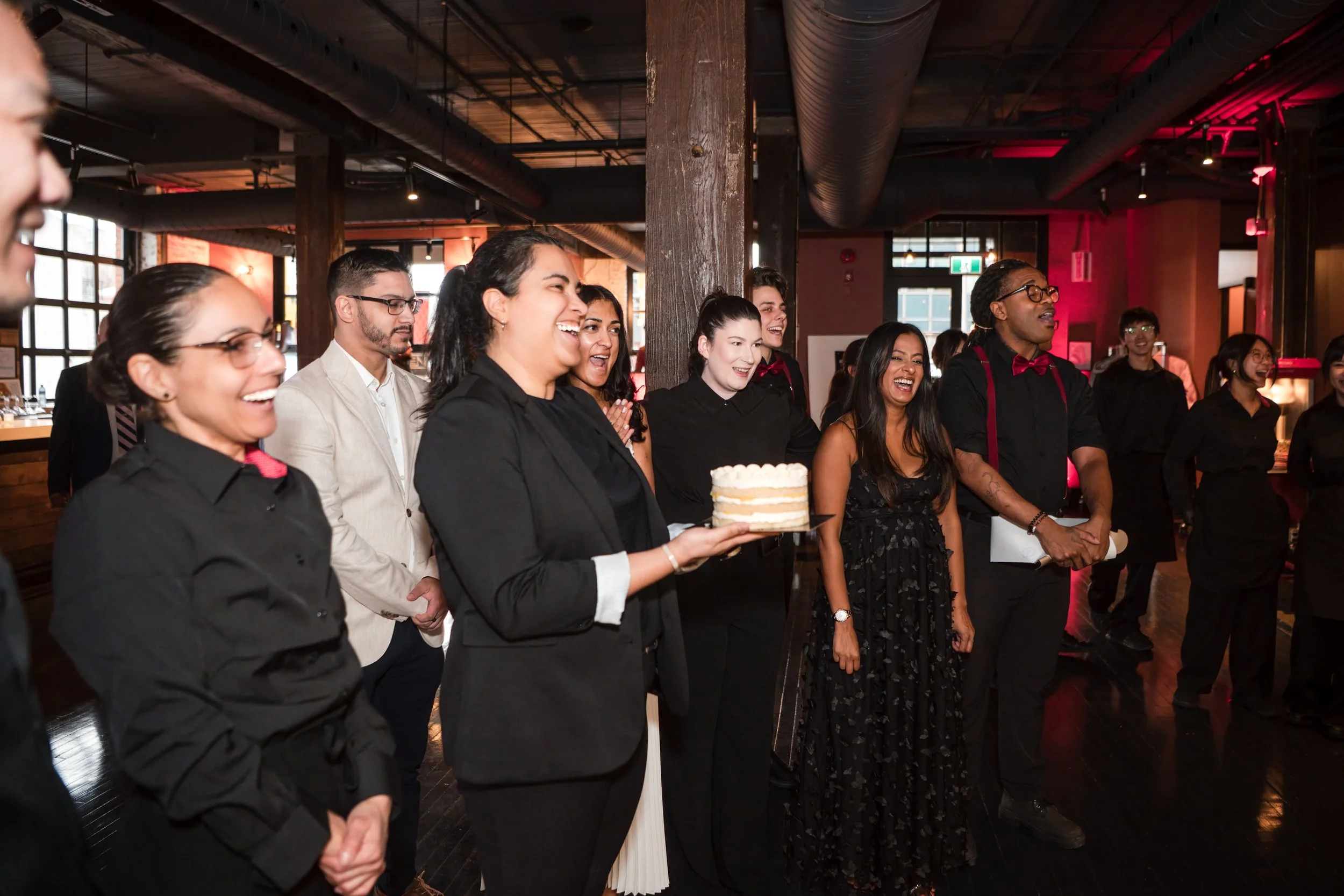 People gathered in a dimly lit indoor space, smiling and laughing, with one woman holding a small layered cake.