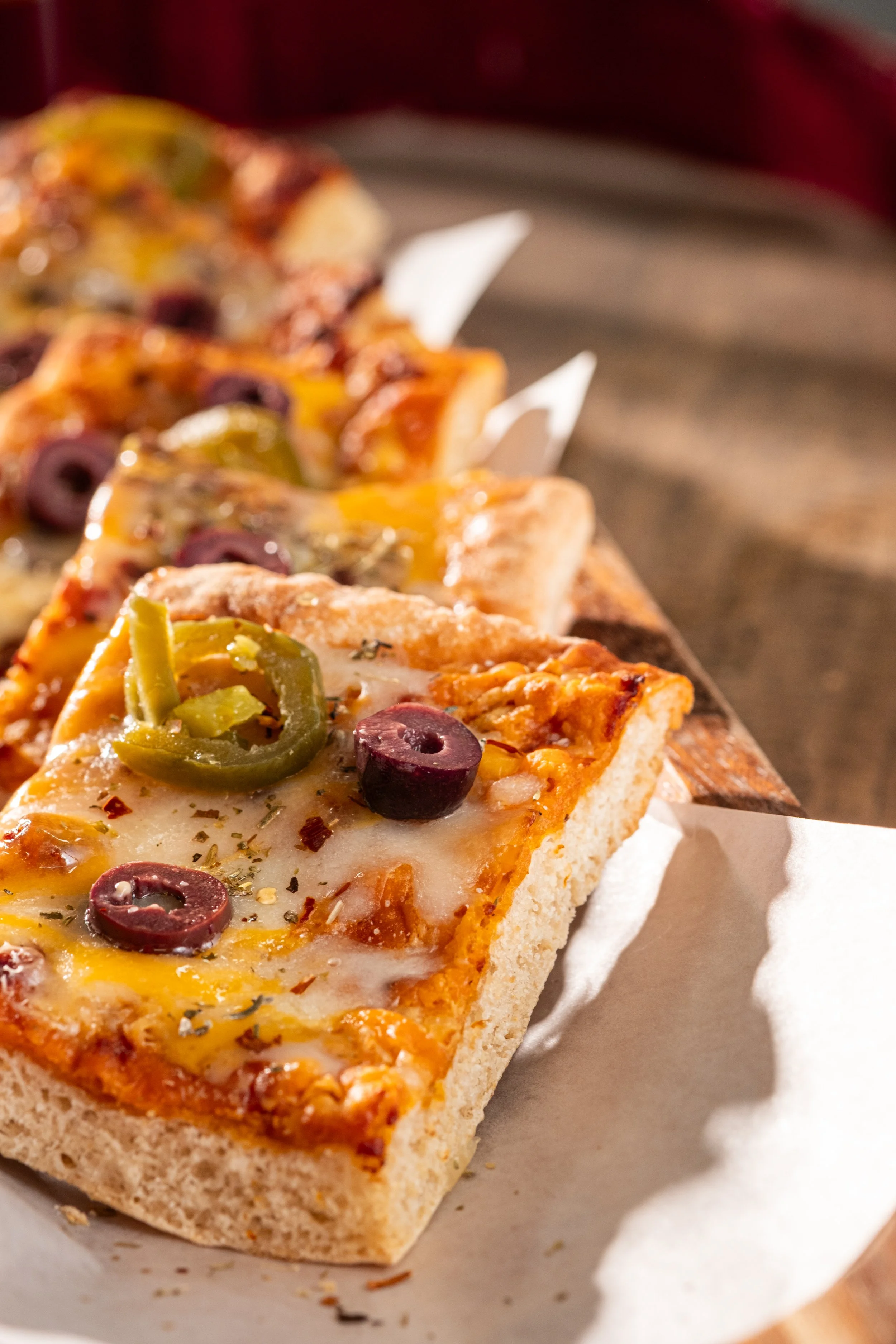 Close-up of a slice of pizza topped with green pepper slices, black olives, and melted cheese on a white paper-lined tray.