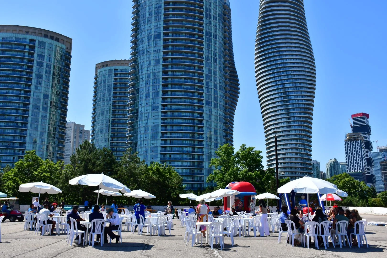 People sitting at tables with white umbrellas in an outdoor setting, with tall modern skyscrapers in the background under a clear blue sky.