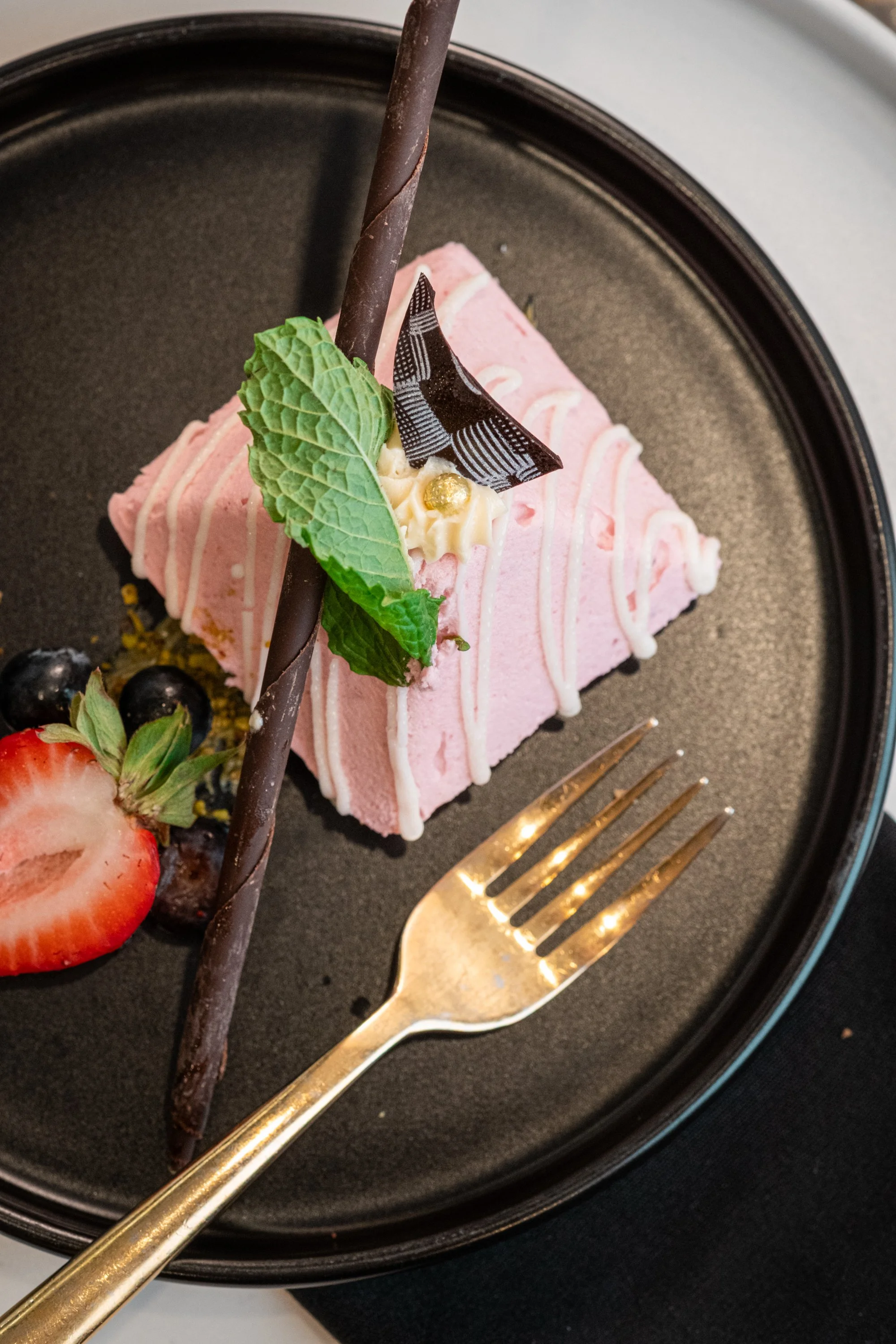 A black plate holding a slice of pink cake with white drizzle and green mint leaf decoration, garnished with chocolate and strawberries, alongside blueberries. A gold fork is placed on the plate.