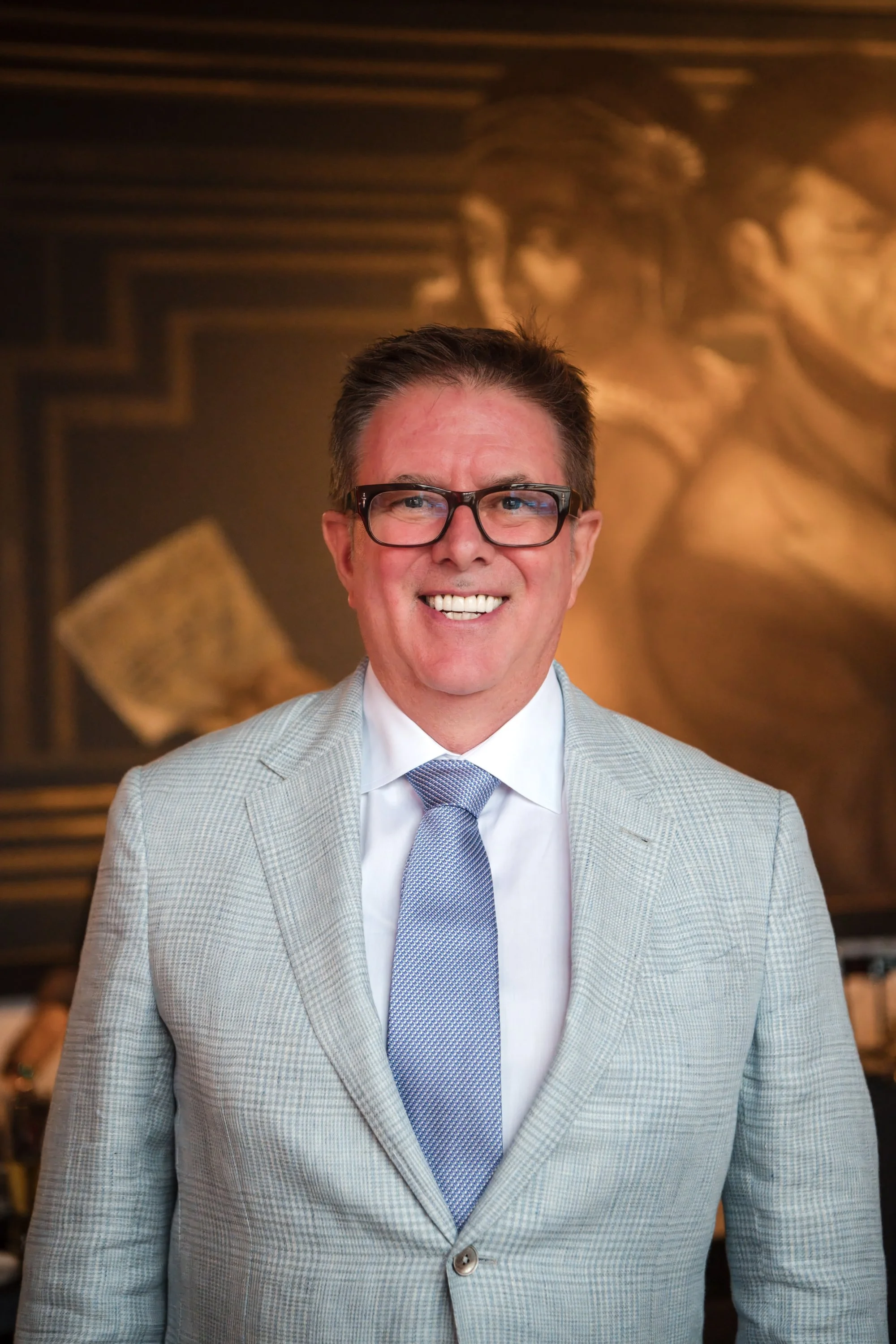 A smiling man in a light gray suit, white shirt, and light blue tie, wearing glasses, standing in front of a gold-colored ornate background.