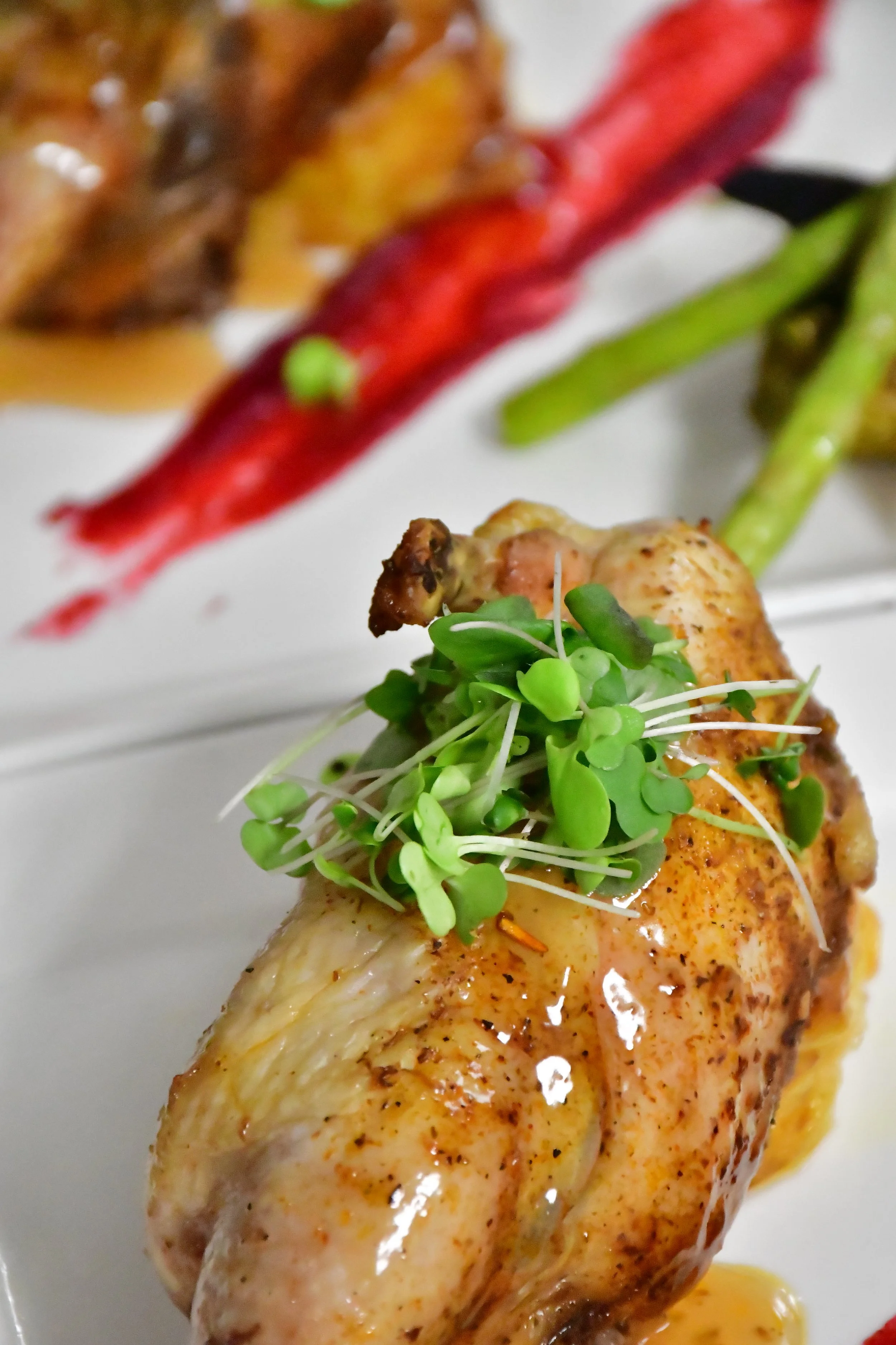 Close-up of a cornish hen leg garnished with microgreens, served with green and red vegetables in the background.