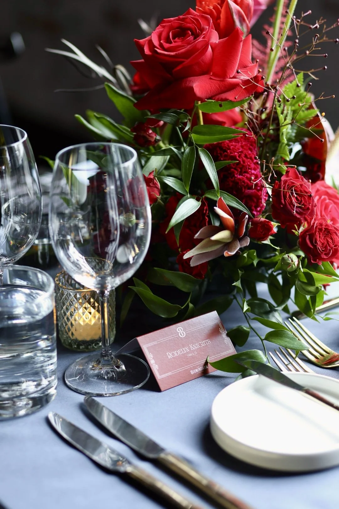 A formal table setting featuring a large floral centerpiece with red roses, greenery, and other flowers, set on a black tablecloth. There are wine glasses, a water glass, a name card, and silverware including a knife, fork, and spoon.