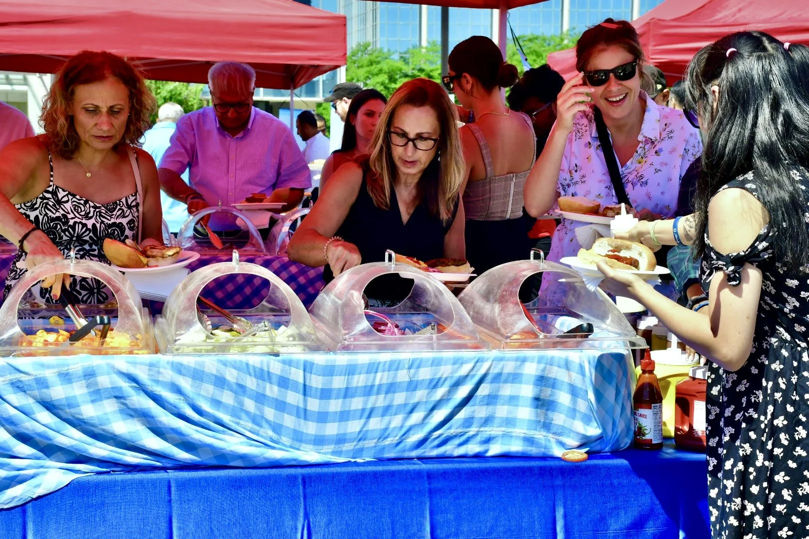 People at a buffet table outdoors, serving themselves hot dogs and condiments on a sunny day.