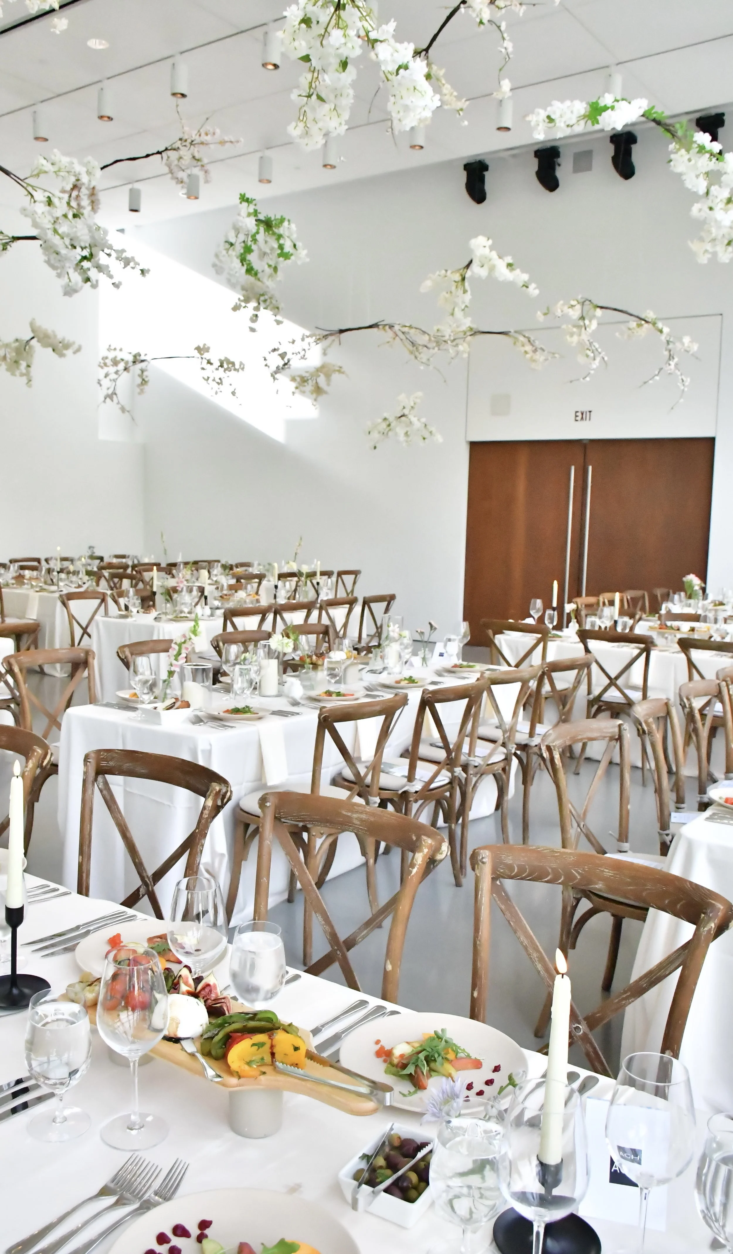 Elegant banquet hall decorated with white linens and wooden chairs, floral centrepieces, candles, and overhead white blossom branches for a wedding or formal event.