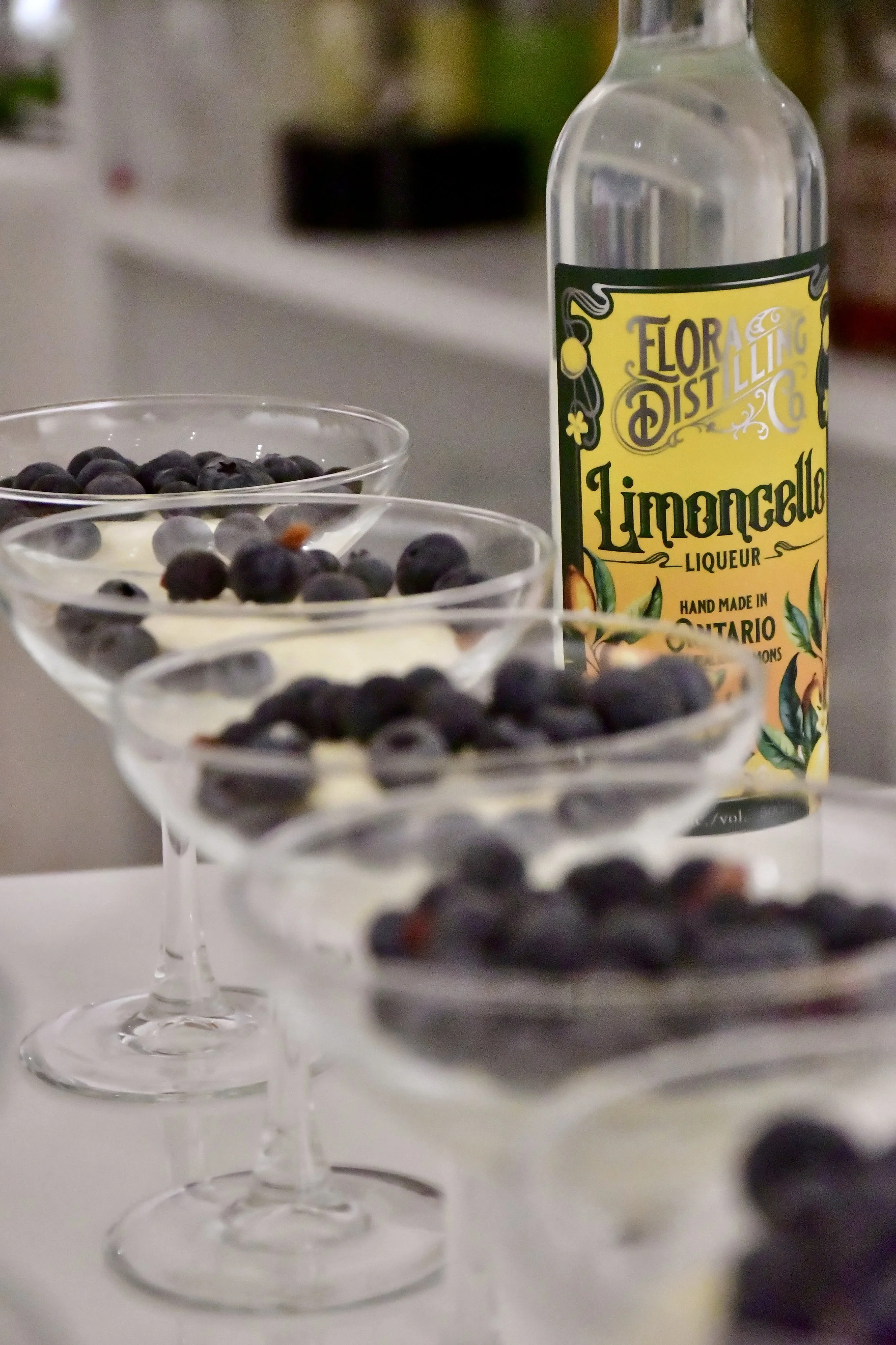 Three cocktail glasses filled with a limoncello mousse dessert topped with blueberries, with a bottle of Elora Distilling Limoncello in the background.