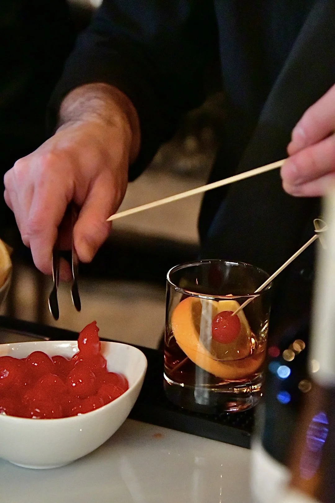 Close-up of a person garnishing a cocktail with cherries and orange slices, with a bowl of cherries nearby.