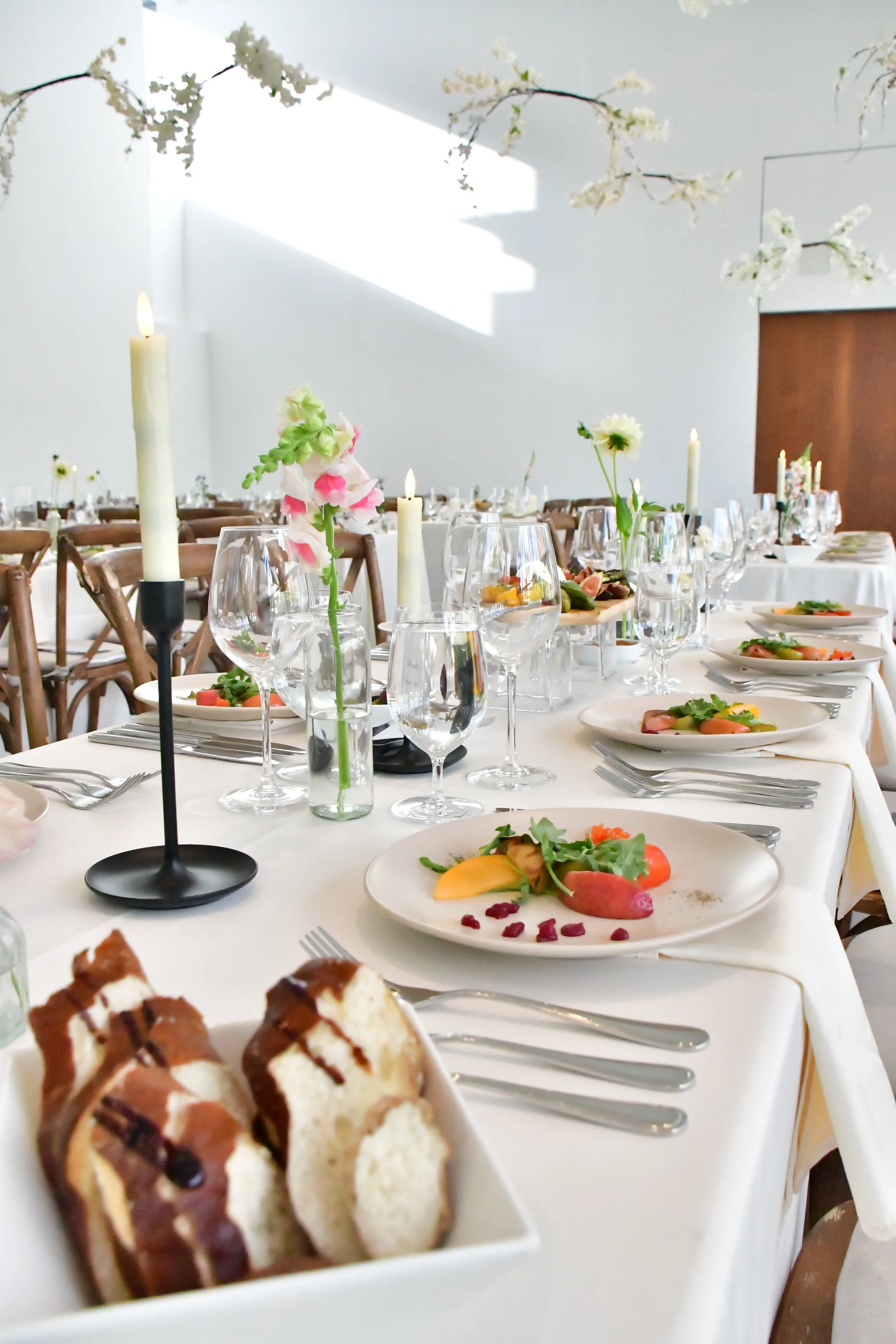 A table set for a formal meal with white tablecloth, wine glasses, plate of food, floral centerpieces, and candles, with a wall and sunlight in the background.
