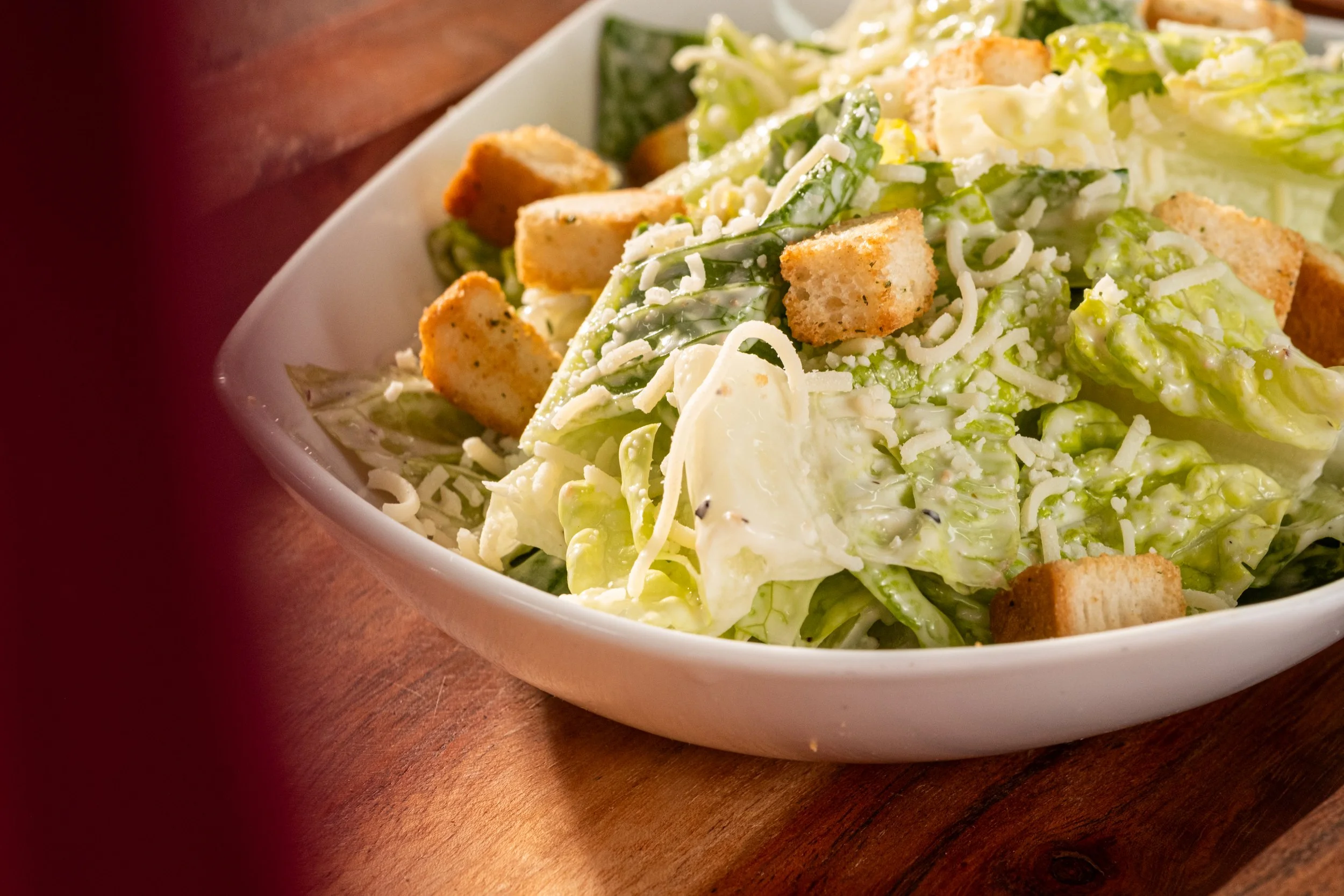 A bowl of Caesar salad with romaine lettuce, croutons, shredded cheese, and dressing on a wooden table.